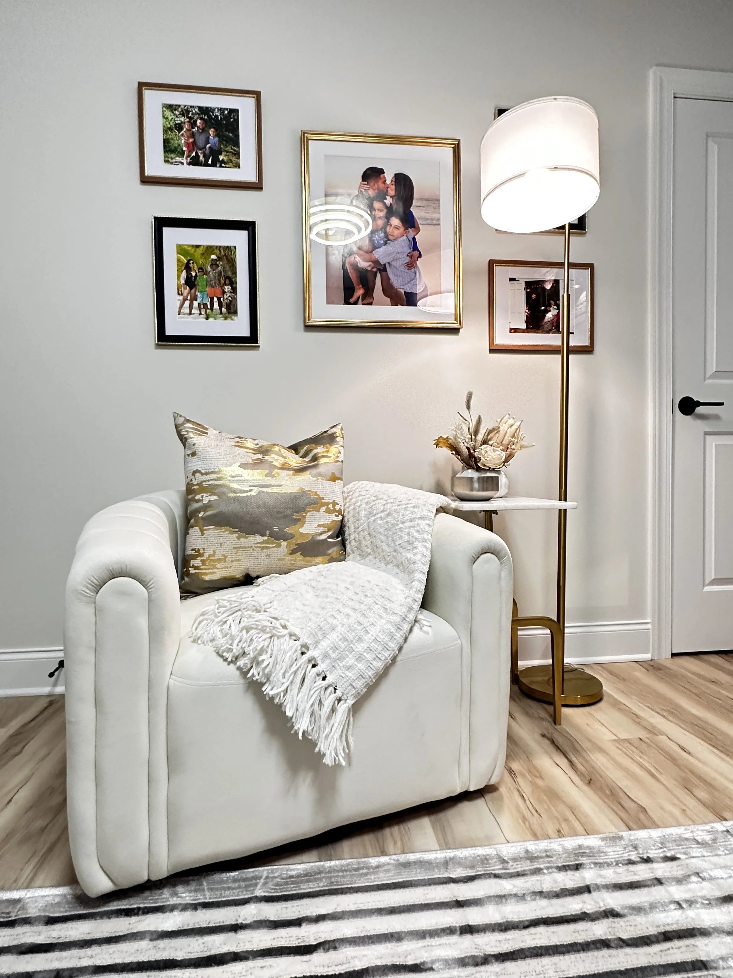Living room with white armchair, decorative pillow, throw blanket, side table with vase of dried flowers, large standing lamp, family photo gallery on the wall, and wooden floor.