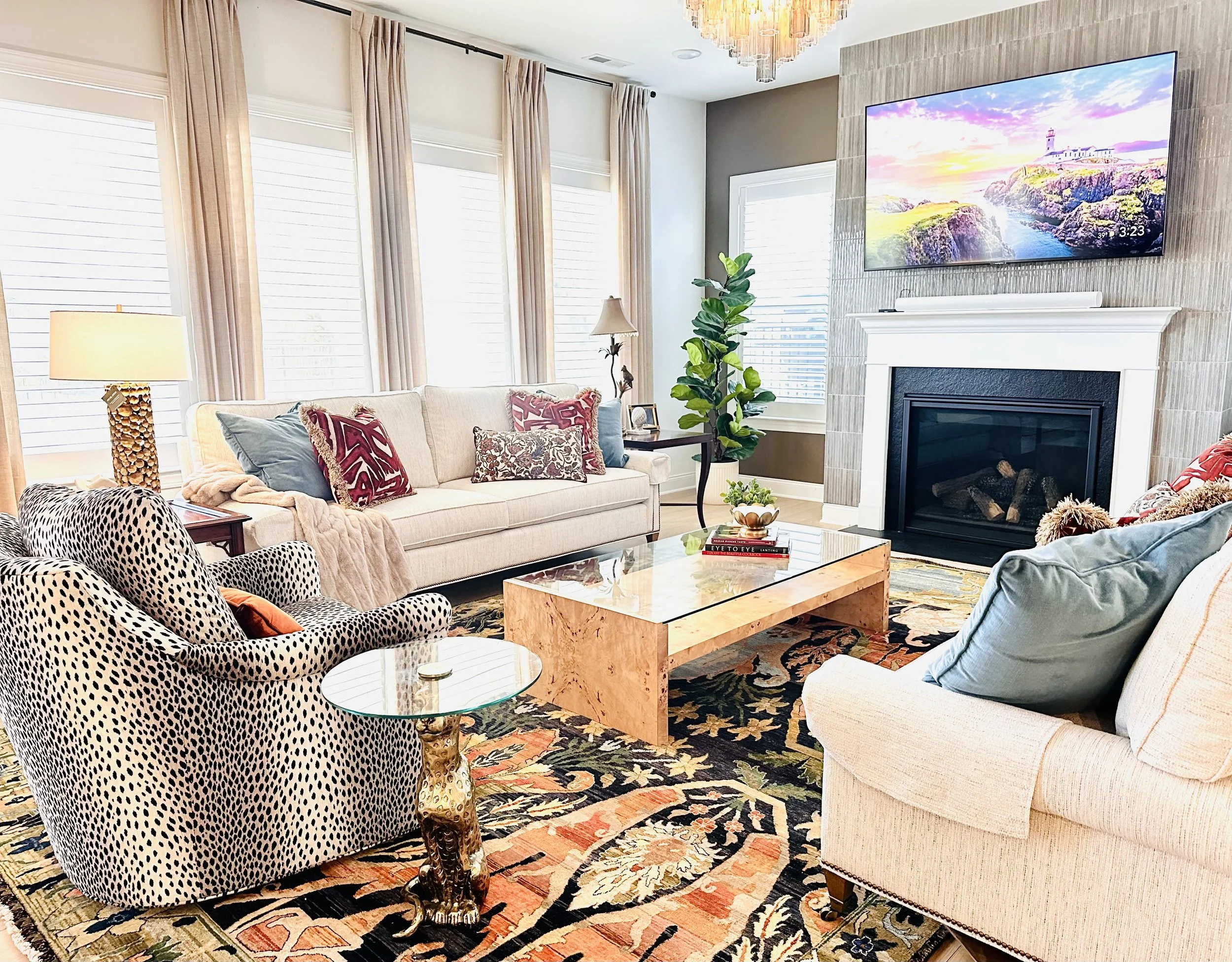 Living room with beige sofas, patterned armchair, colorful cushions, a marble-topped coffee table, a fireplace, a large TV, and windows with beige curtains.
