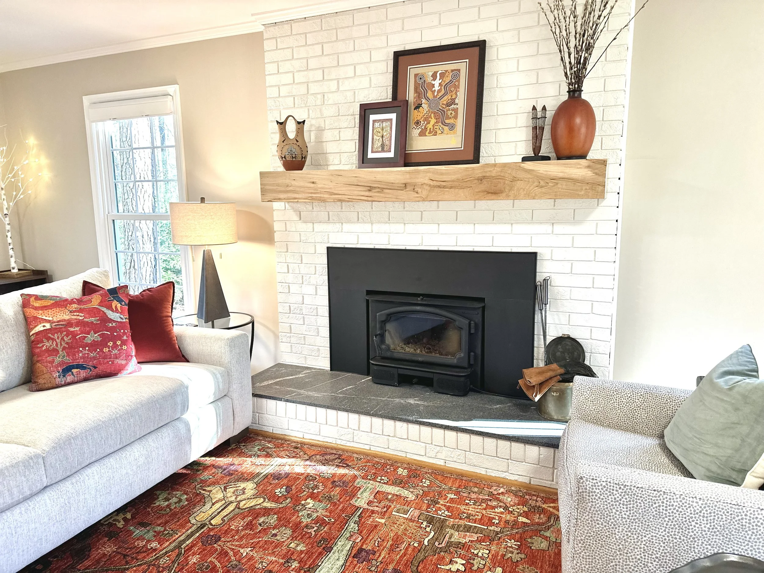 Living room with a fireplace, white brick wall, wooden mantel decorated with framed artwork, vases, and branches. Beige sofa with colorful pillows, side table with a lamp, window with greenery outside, and a patterned rug.