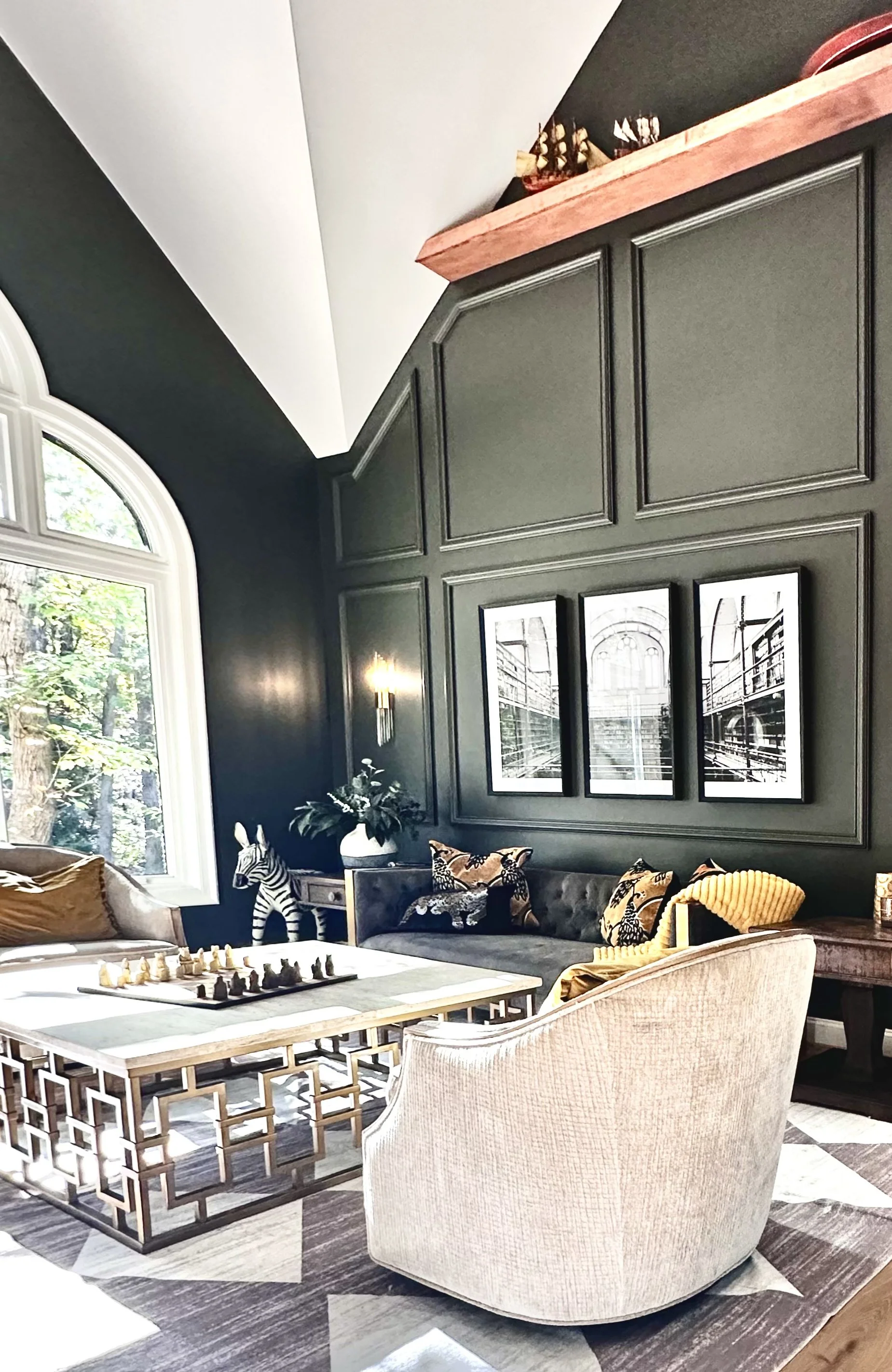 Stylish living room with dark green wall, large arched white window showing trees outside, a beige armchair, a dark gray velvet sofa with patterned pillows, a white coffee table with chess, and a wooden side table, with decorative sailing ship models on a shelf.