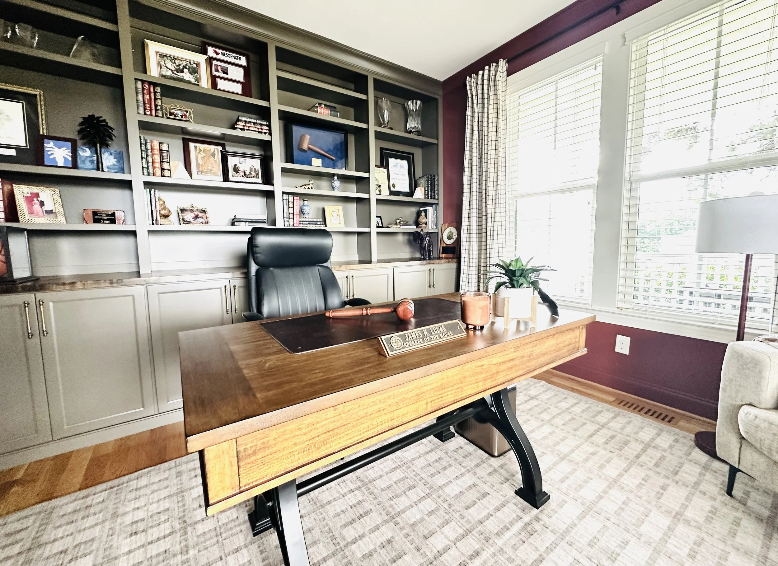 A modern office with a wooden desk, black leather chair, large window with blinds, and built-in shelves filled with books and decorative items.