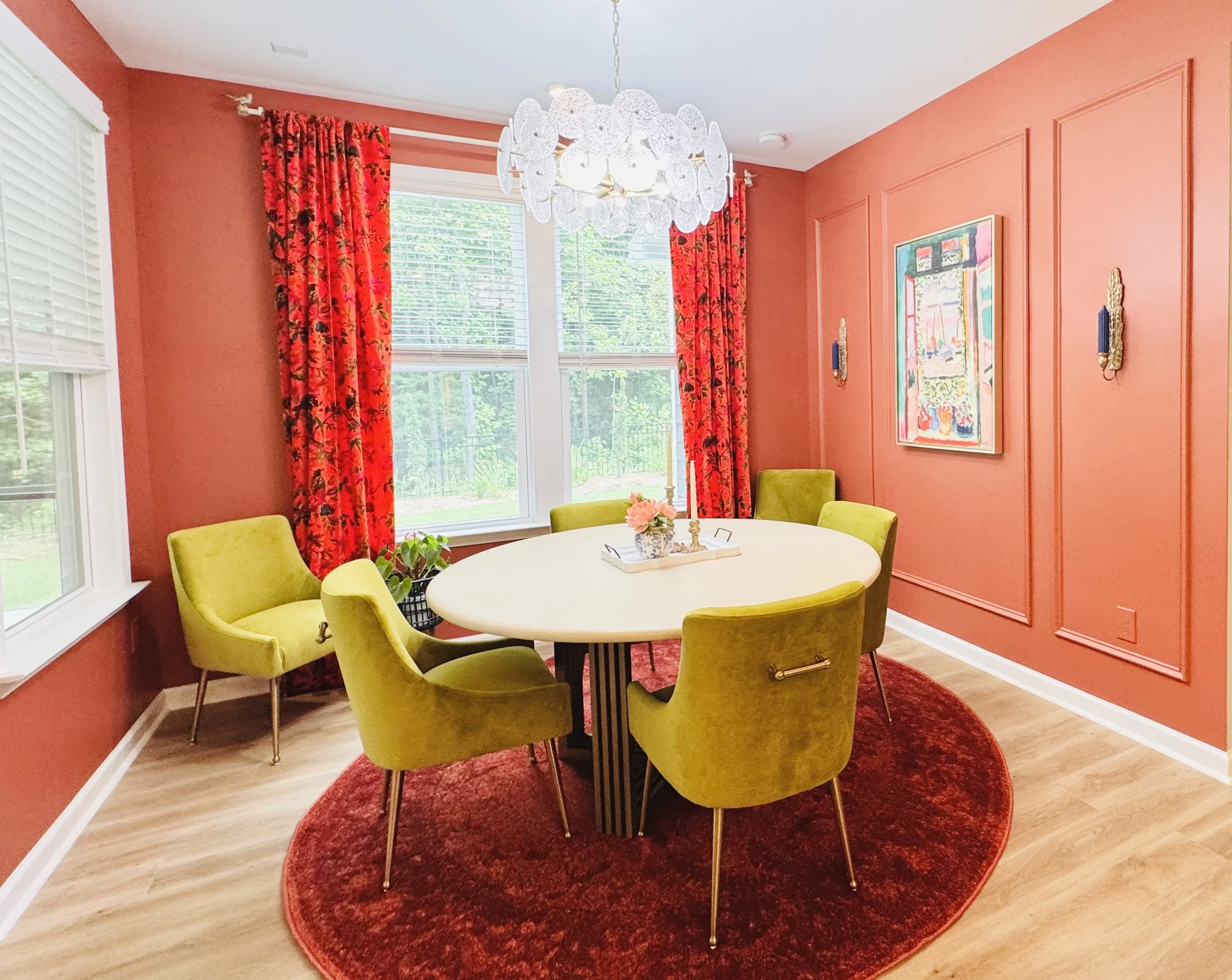 A bright dining room with coral walls, red floral curtains, and a white oval table surrounded by six green velvet chairs. A white chandelier hangs above the table, and a painting decorates the wall. The room has large windows with white blinds revealing a view of trees outside, and a red round rug is beneath the table.