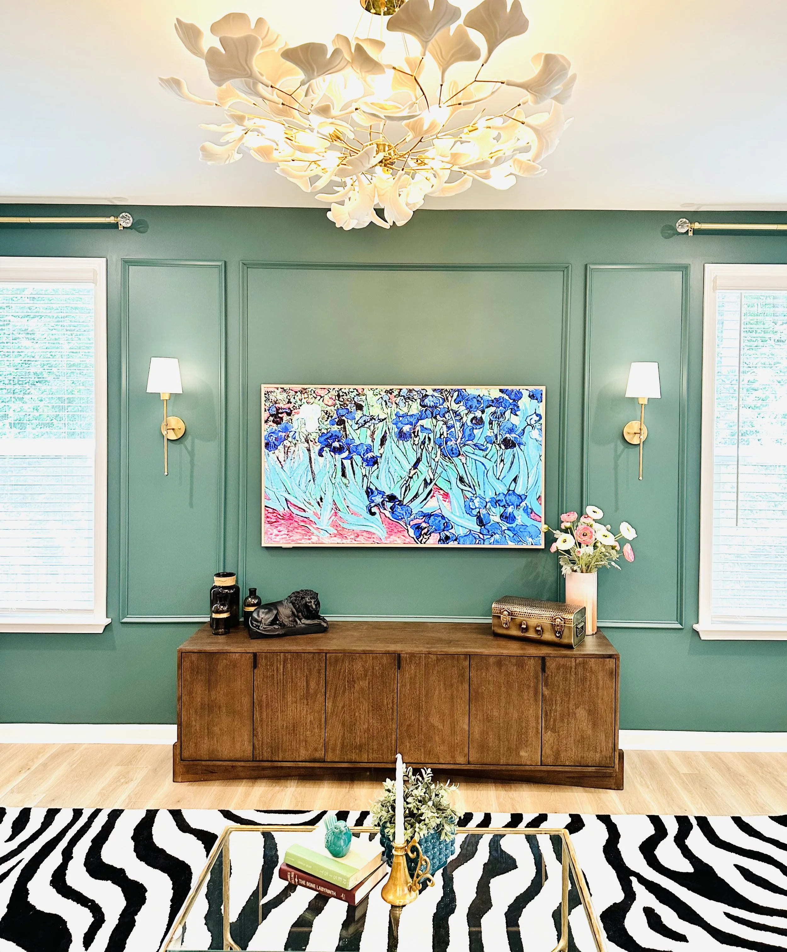 Living room with green wall, two windows with blinds, and artwork of blue irises. Includes a wooden cabinet with black decor, a white vase with pink and white flowers, and a gold decorative radio. A zebra-patterned rug and glass coffee table with books and a candle centerpiece.
