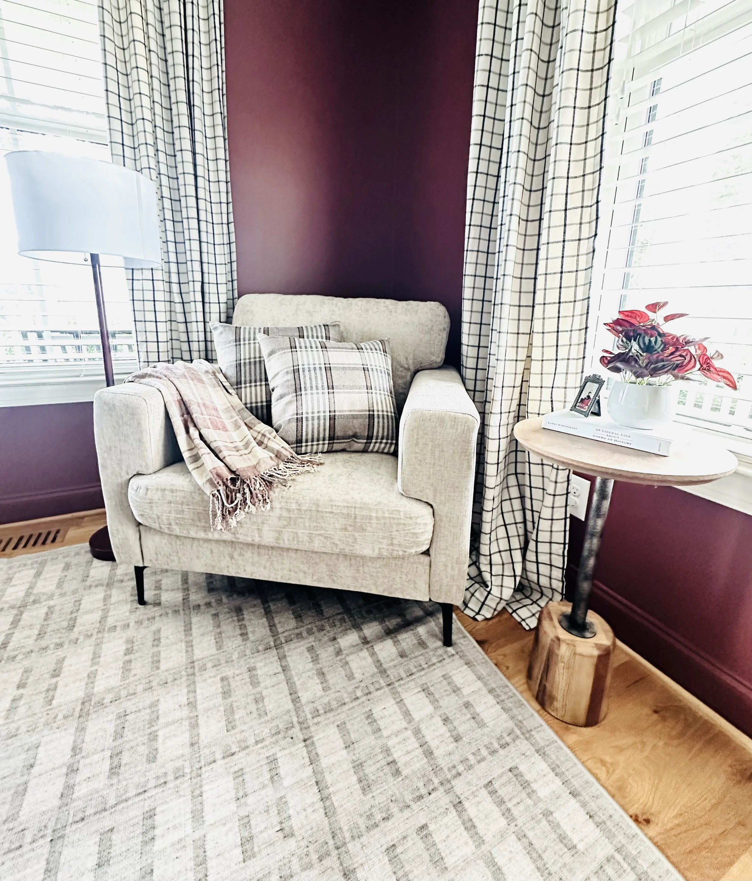 A cozy corner with a beige armchair, plaid cushions, a pink and beige plaid throw, a side table with a potted plant, books, and framed photo, and a window with white blinds and checkered curtains, all against a burgundy accent wall.