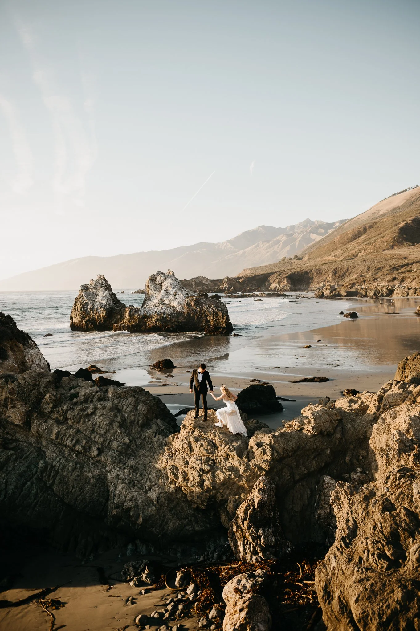 A couple dressed in wedding attire on rocky beach, with a man helping a woman on her knees.