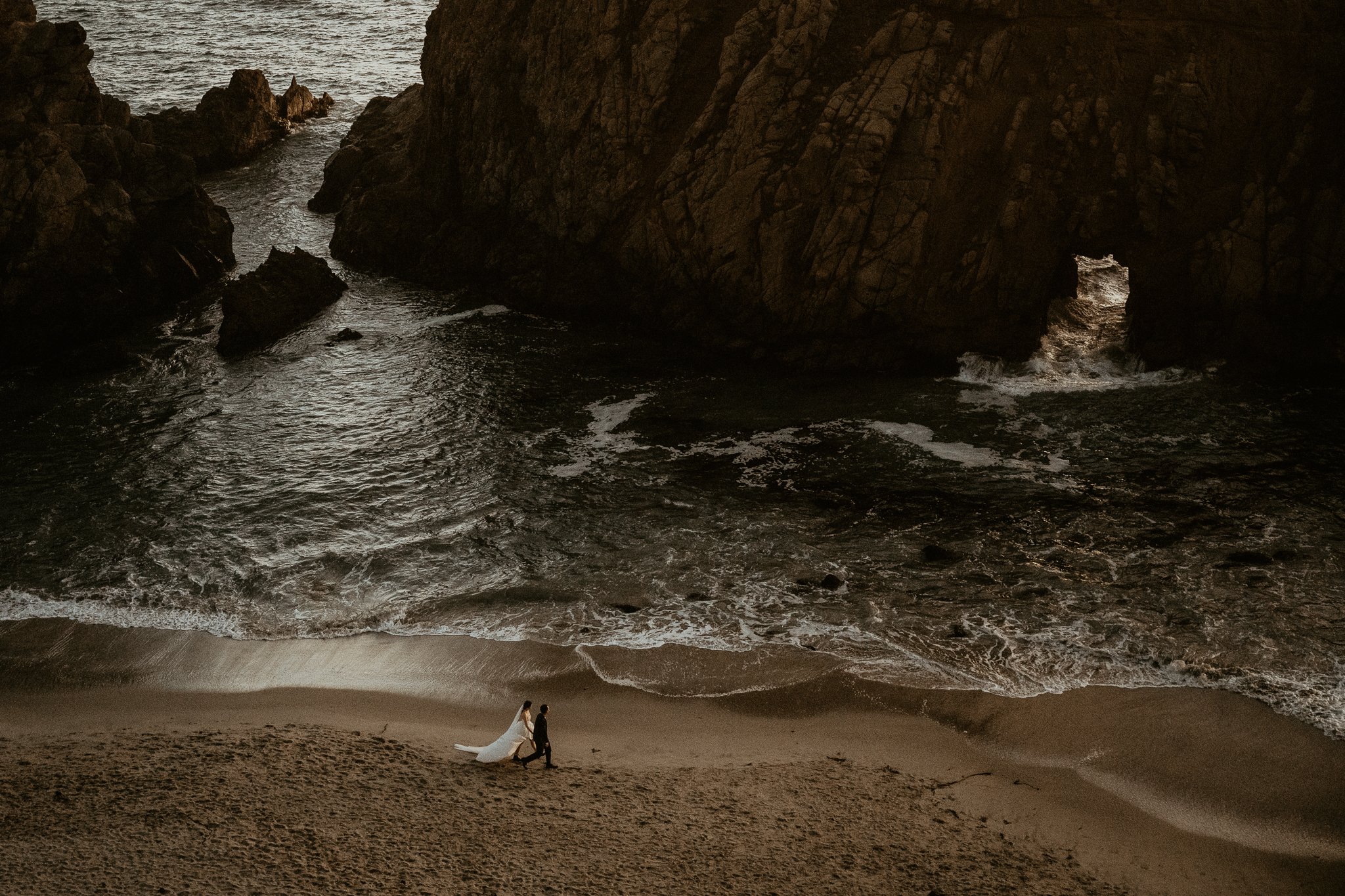 A bride and groom walk hand in hand along a beach near a large rocky cliff with a natural arch, during sunset or dusk.