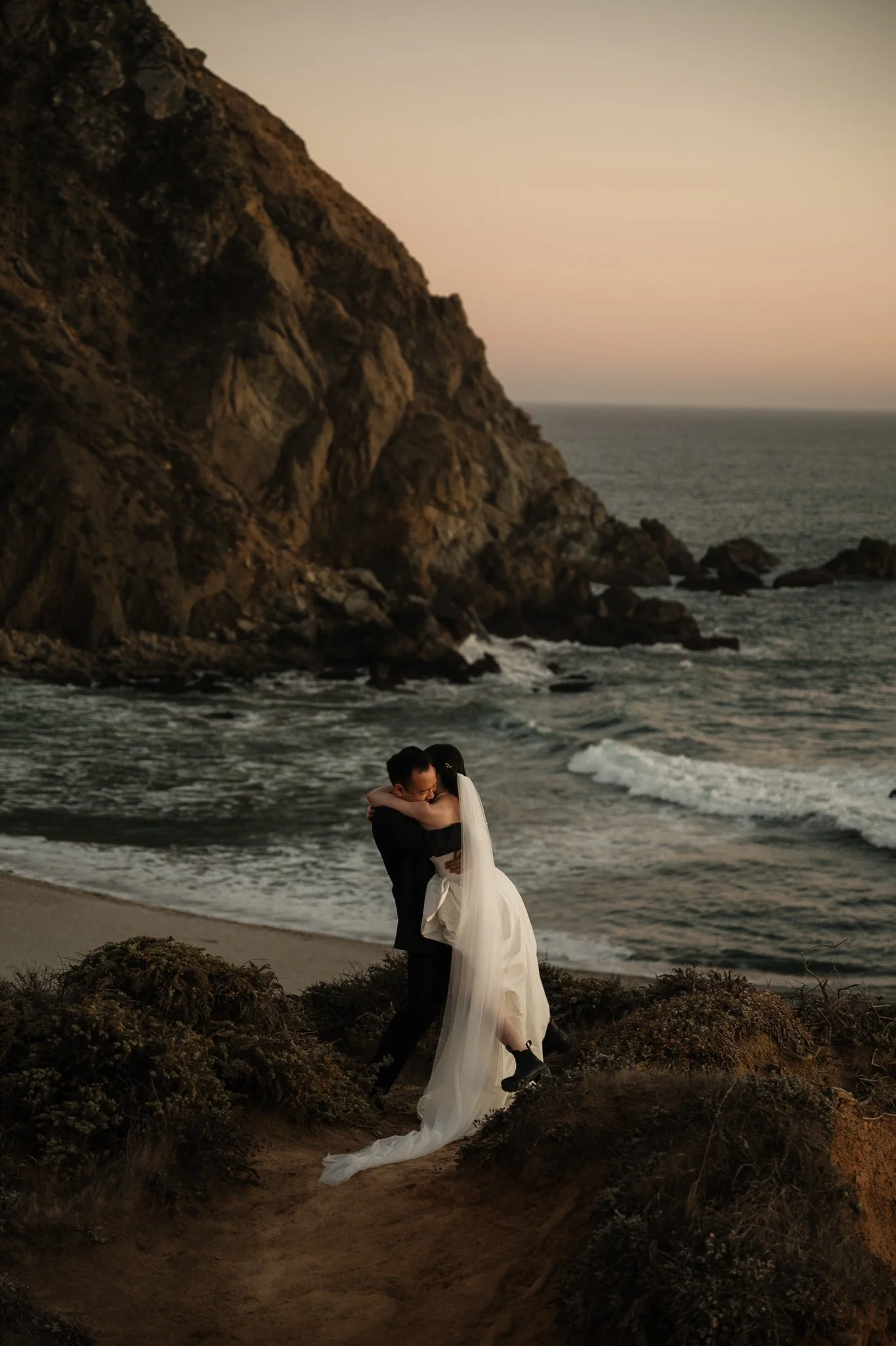 A couple in wedding attire embracing on a beach at sunset, with a rocky cliff in the background.
