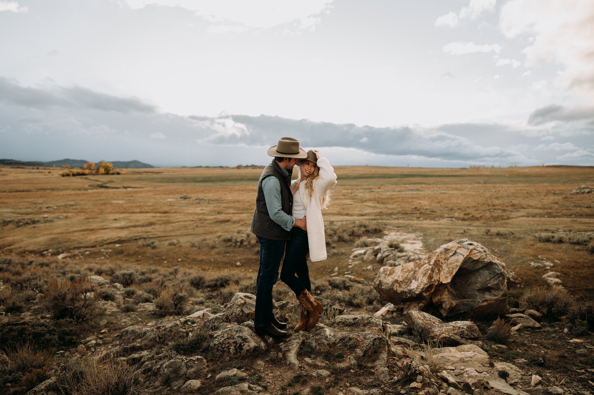 Wedding couple in casual dress in a big open grassy field standing close together bride is hold her cowboy hat at Brush Creek Ranch, Wyoming