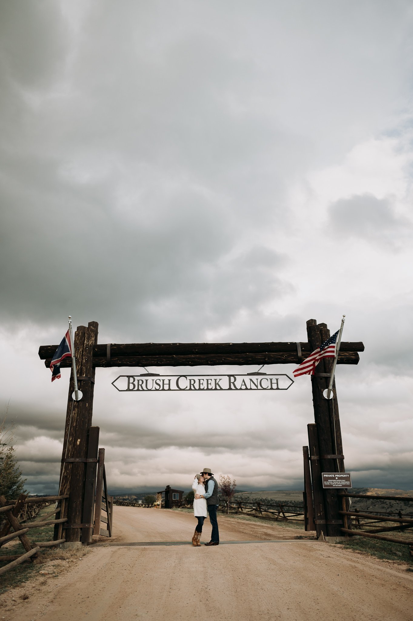 Brush Creek Ranch Wyoming couple hugging in drive under the entry sign with a cloudy grey sky 