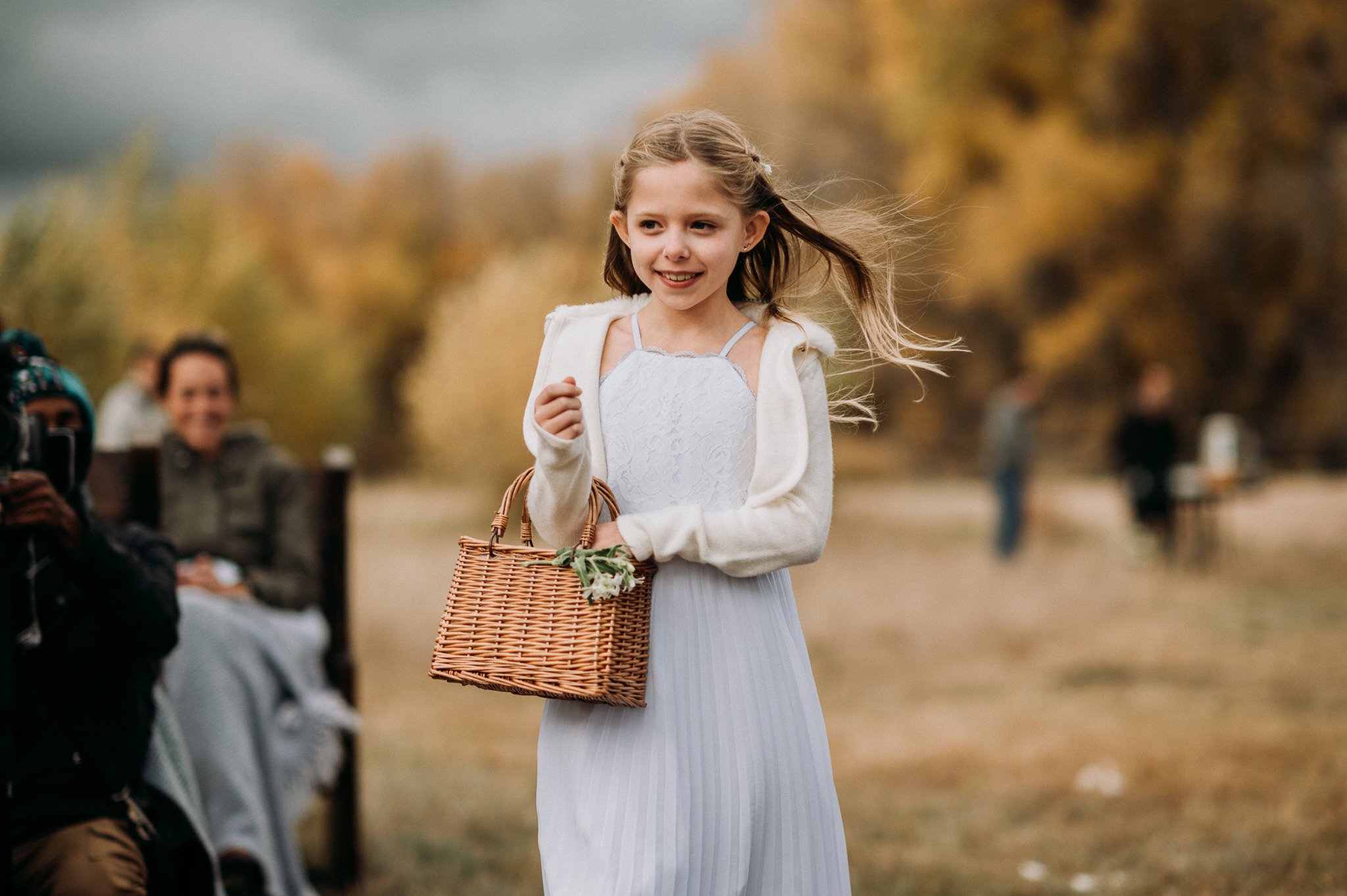 young flower girl in white dress walking down the isle outside in a grassy field with the orange and yellow trees of autumn in the background.