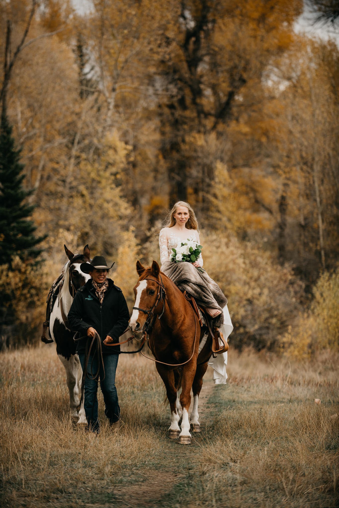 Bride in wedding dress riding side saddle on horse to ceremony site in grassy field with autumn colors in the trees in back ground. 