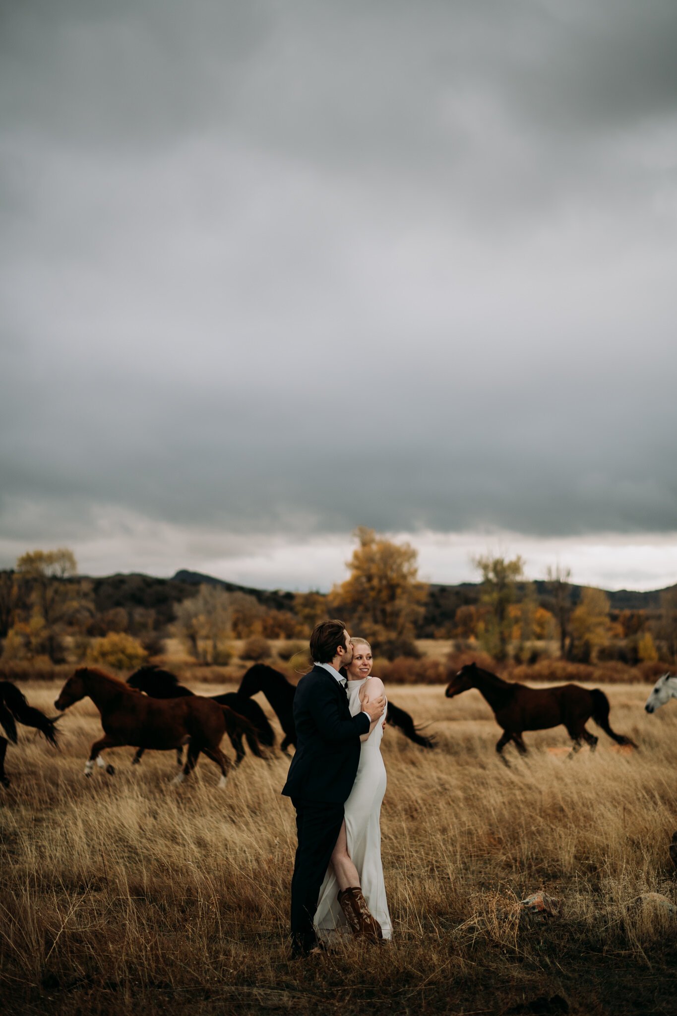 Bride and groom in wedding attire in grassy field with hoses running in the background and the big Wyoming grey sky.