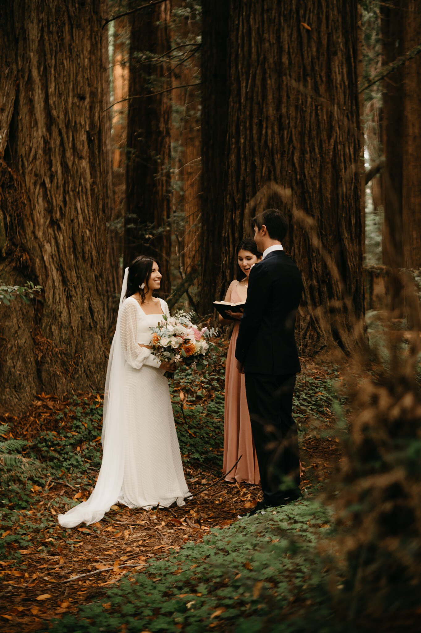 Bride and groom in forest ceremony saying vows in front of officiant, with tall redwood trees in background.
