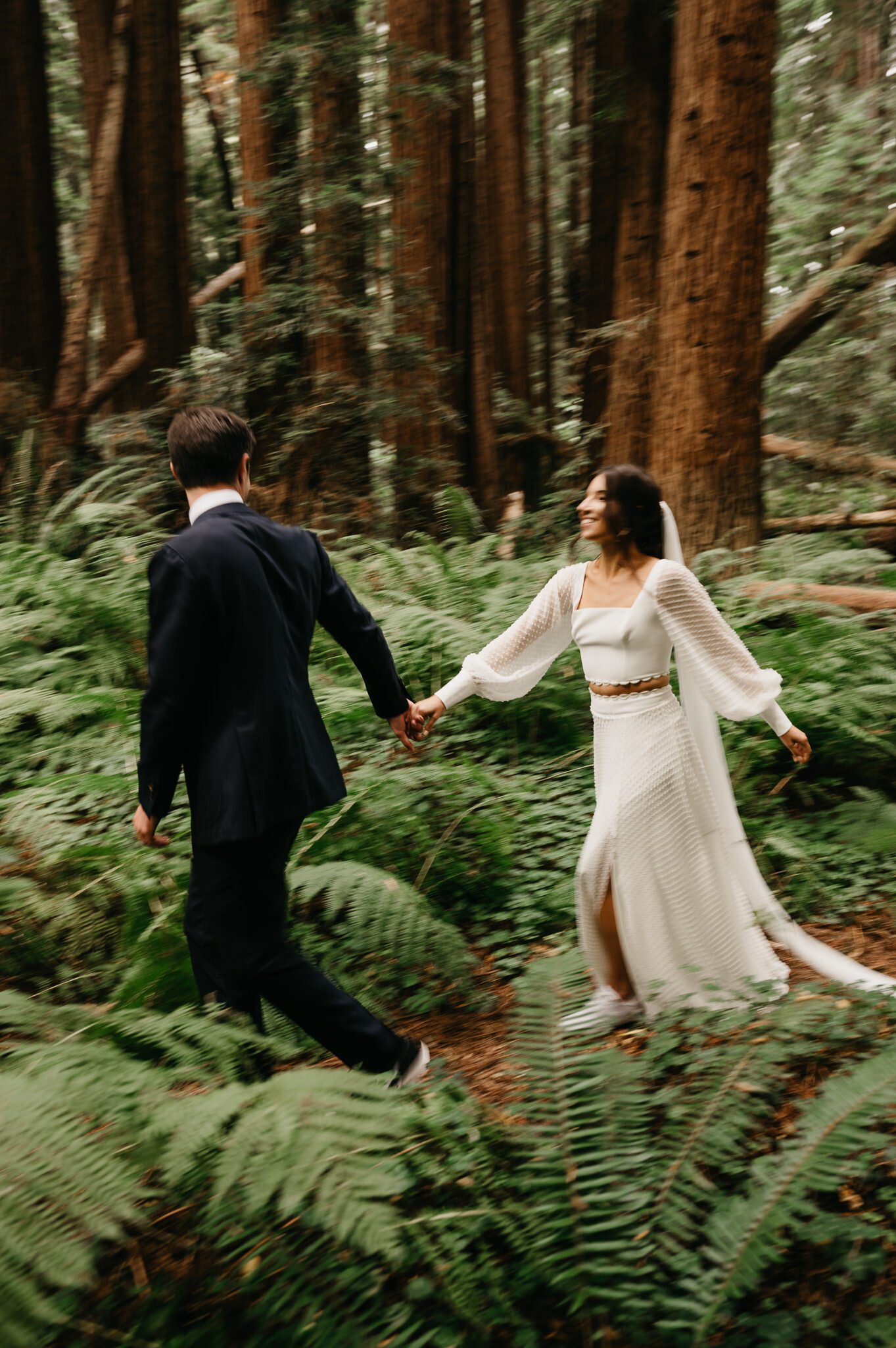 Newly married couple holding hands walking through ferns after their forest elopement.