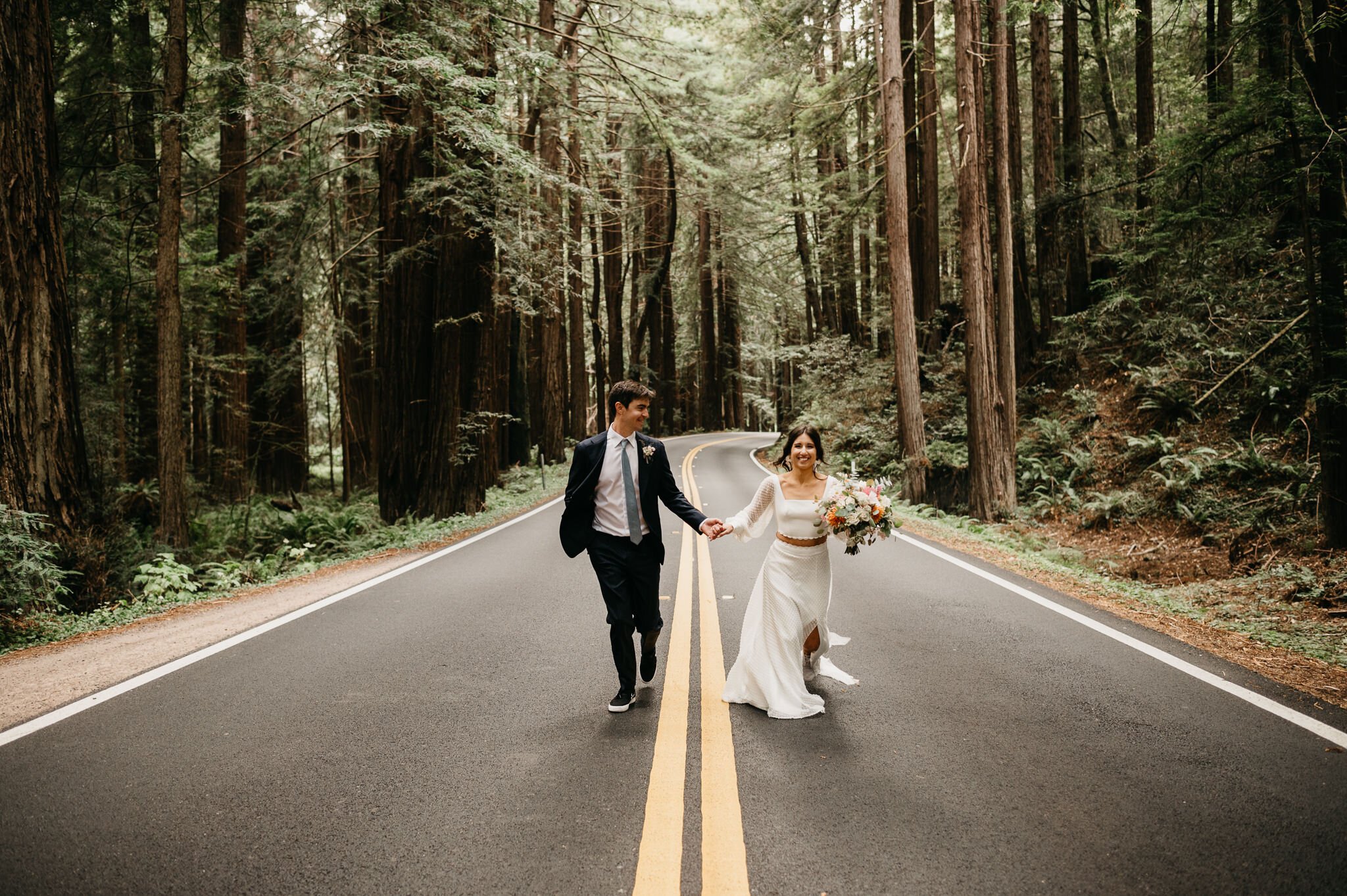 Bride and groom in wedding attire holding hands walking down middle of the road, with redwood trees on both sides of them after their forest ceremony.
