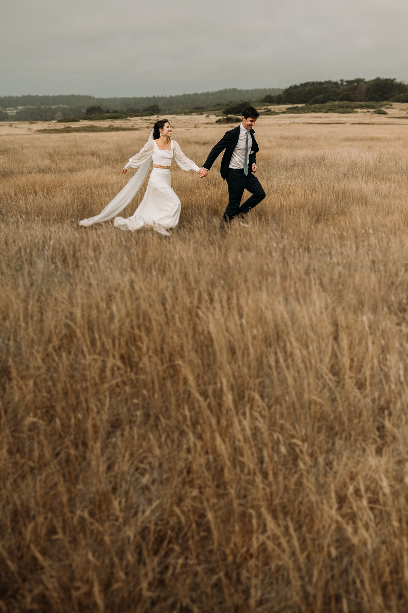 Bride and groom in wedding attire running through grassy field hand and hand on the Mendocino coast.