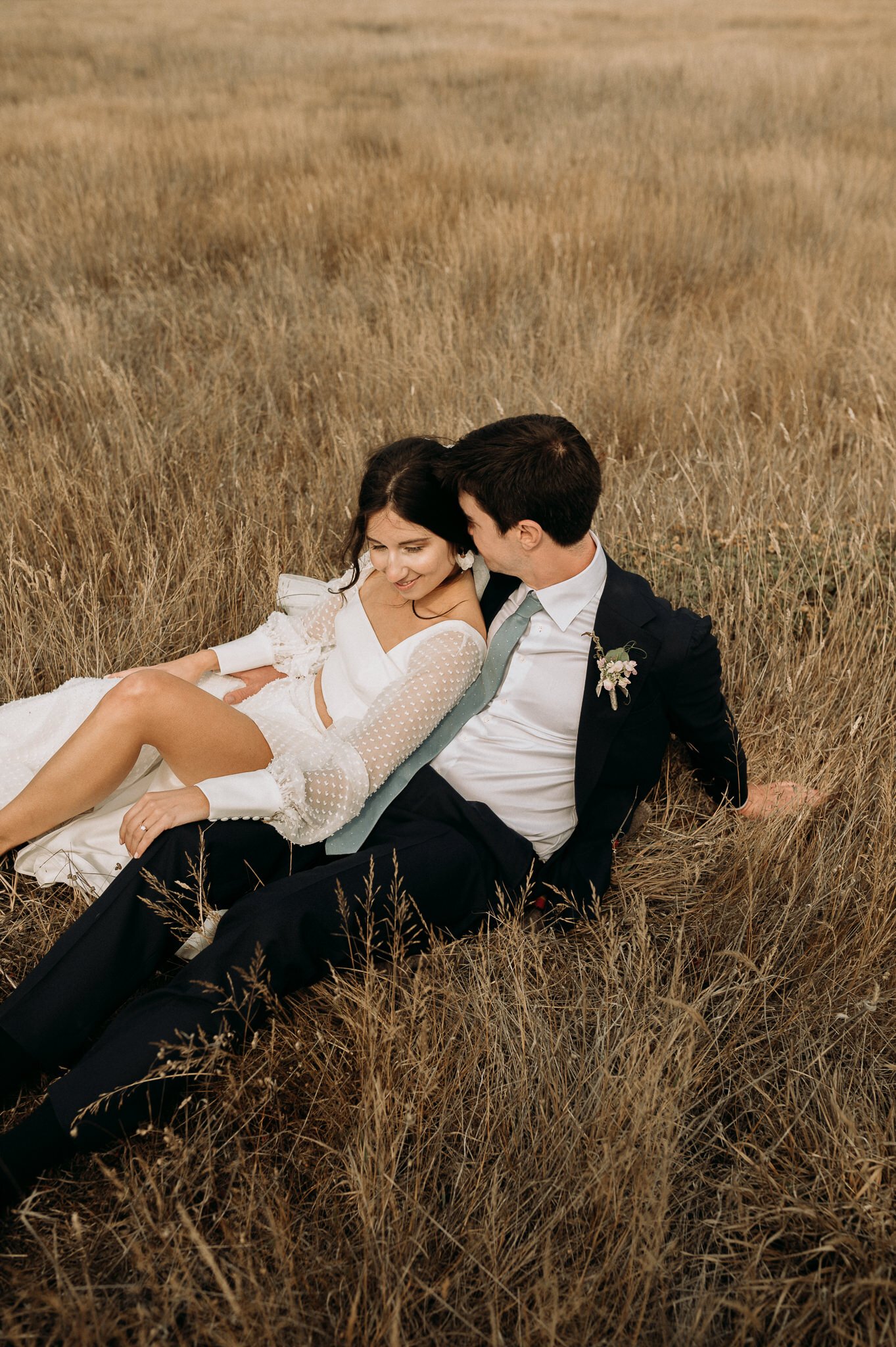 newly married couple laying in grassy field along the coast on California, bride is laying against grooms chest, she is looking out at the Pacific Ocean and he is looking at her. 