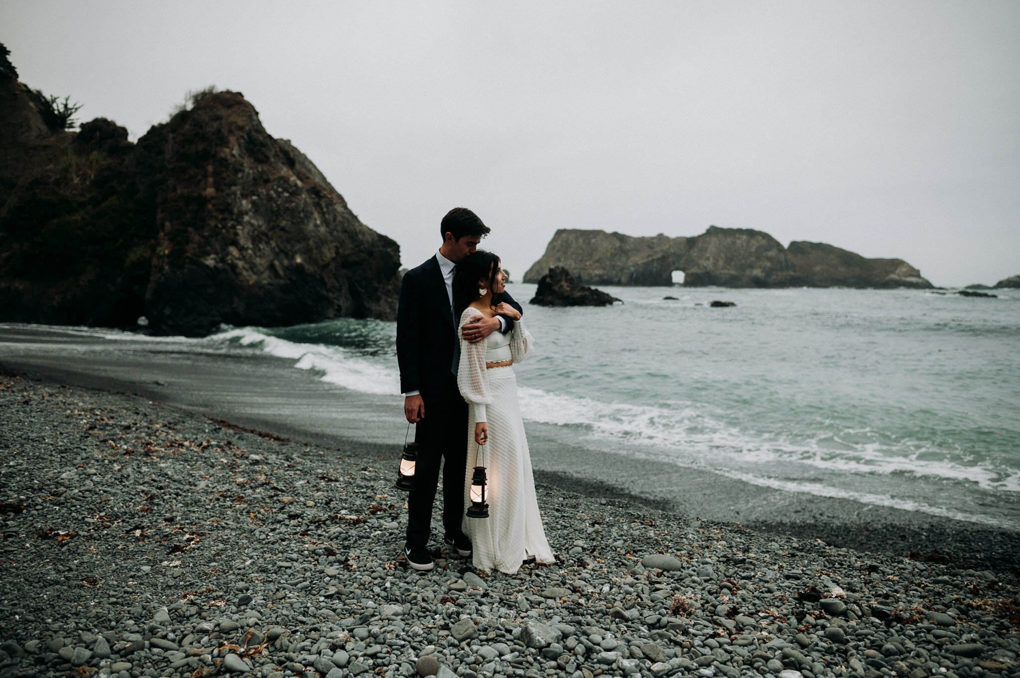 newly married couple in wedding attire embracing with groom standing behind bride both are hold lanterns on the beach in Mendocino.
