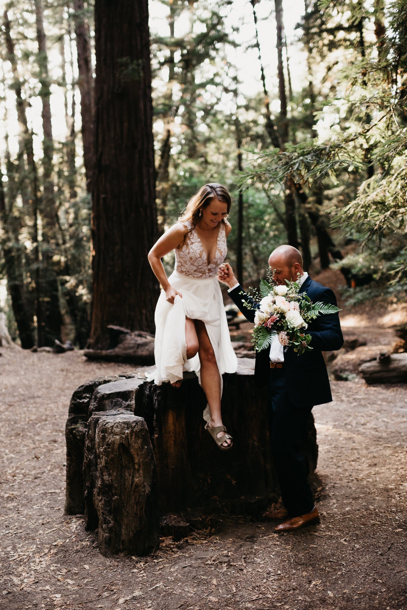 Groom helping bride down from tree stump in Forest Big Sur