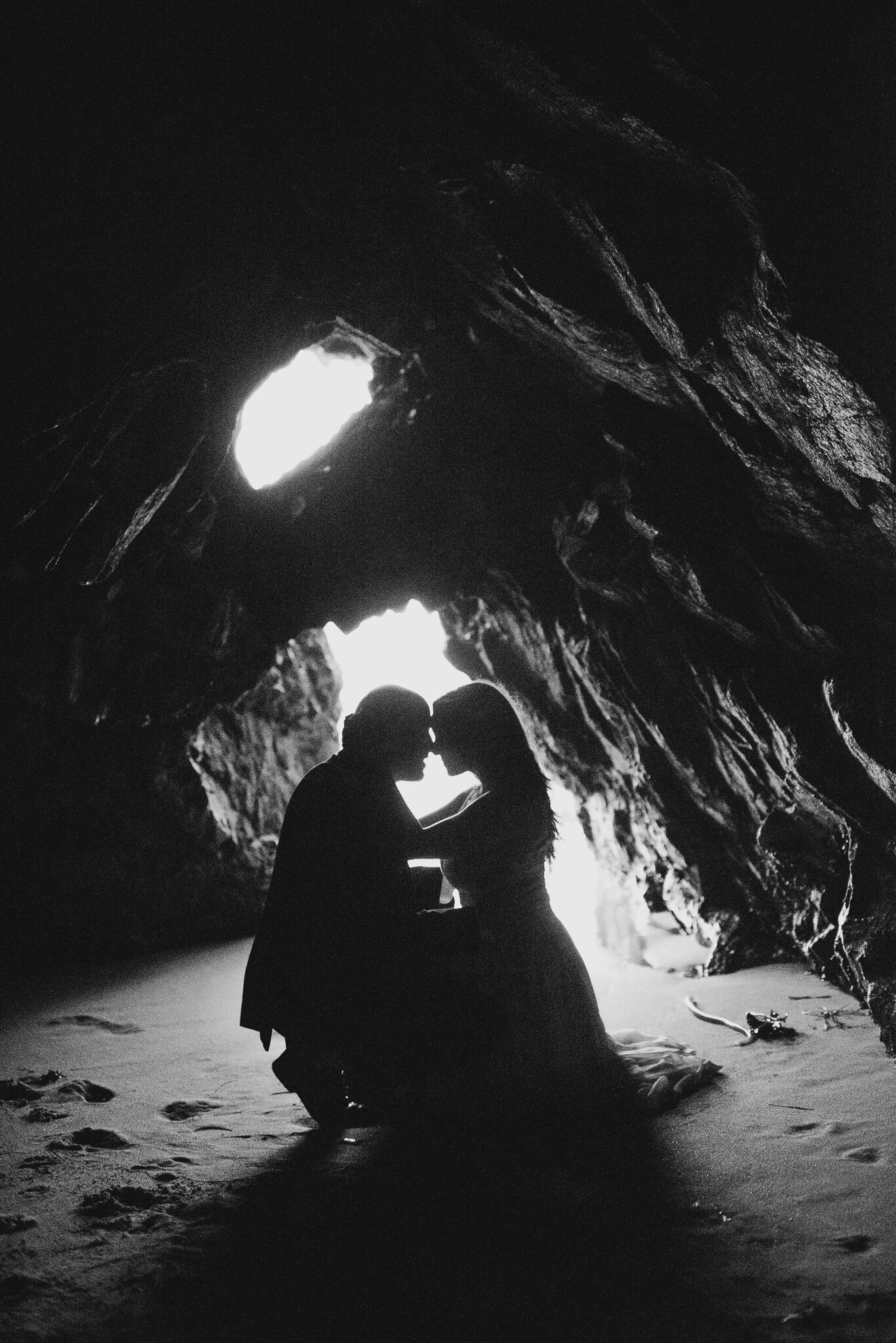 black and white of bride and groom in ocean cave kneeling with foreheads together her arms on his neck with light shining through behind them
