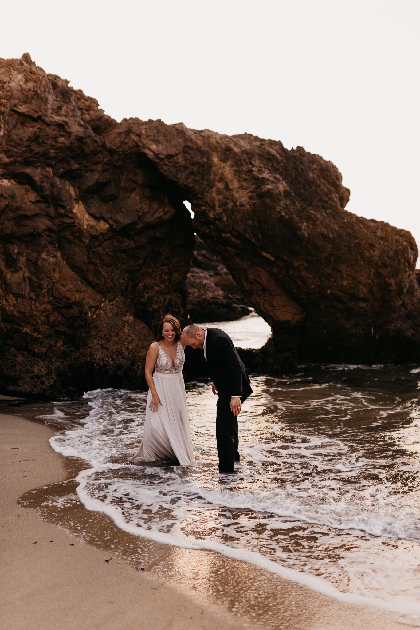 Ventana Elopement Bride and groom in wedding clothing with a wave rolling over their feet with large rock in background