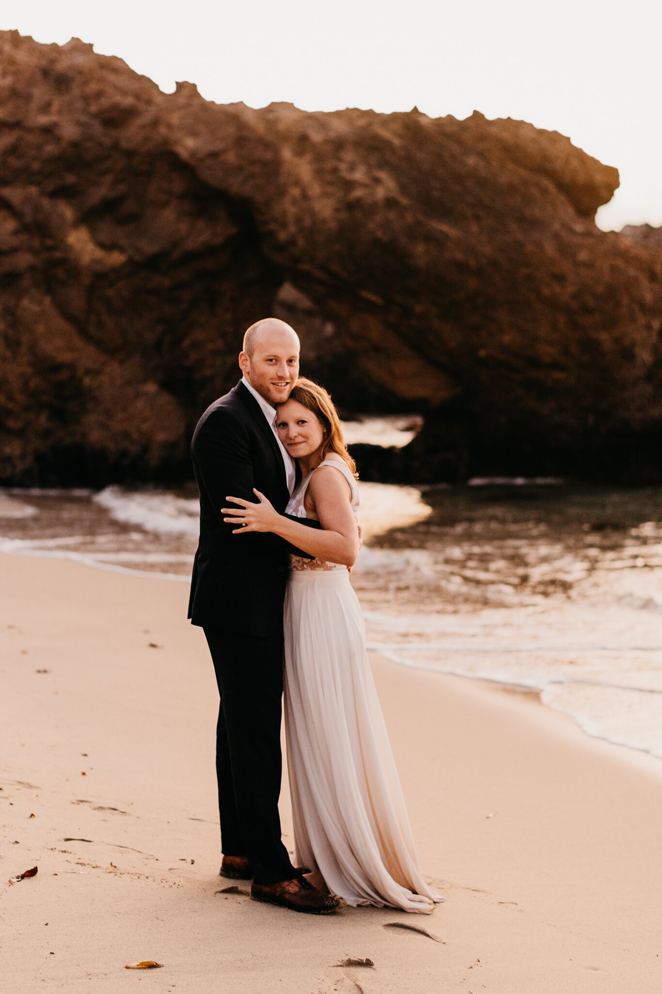 Big Sur Elopement couple in wedding attire on beach embracing,  large rock with key hole in background