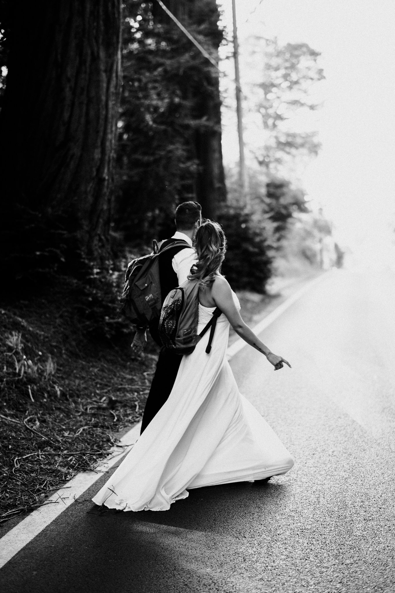 newly married couple walking across road dressed wedding attire with backpacks and hiking boots California Hwy 1
