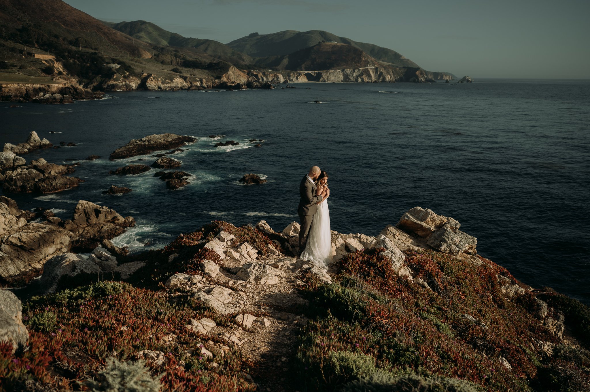 Bride and groom posing cliffside with Bixby Creek bridge and pacific ocean in background