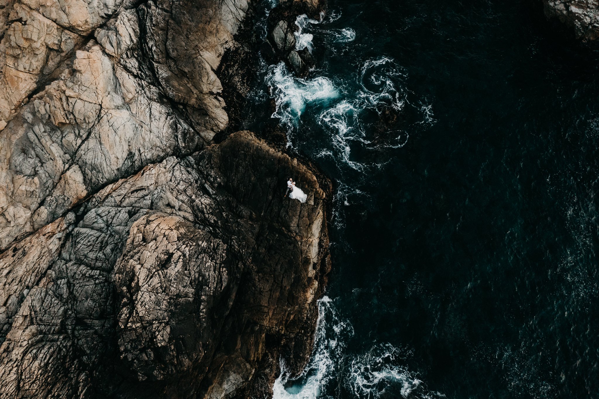 Drone photo of newly married couple laying on cliff with pacific ocean surrounding them in Big Sur