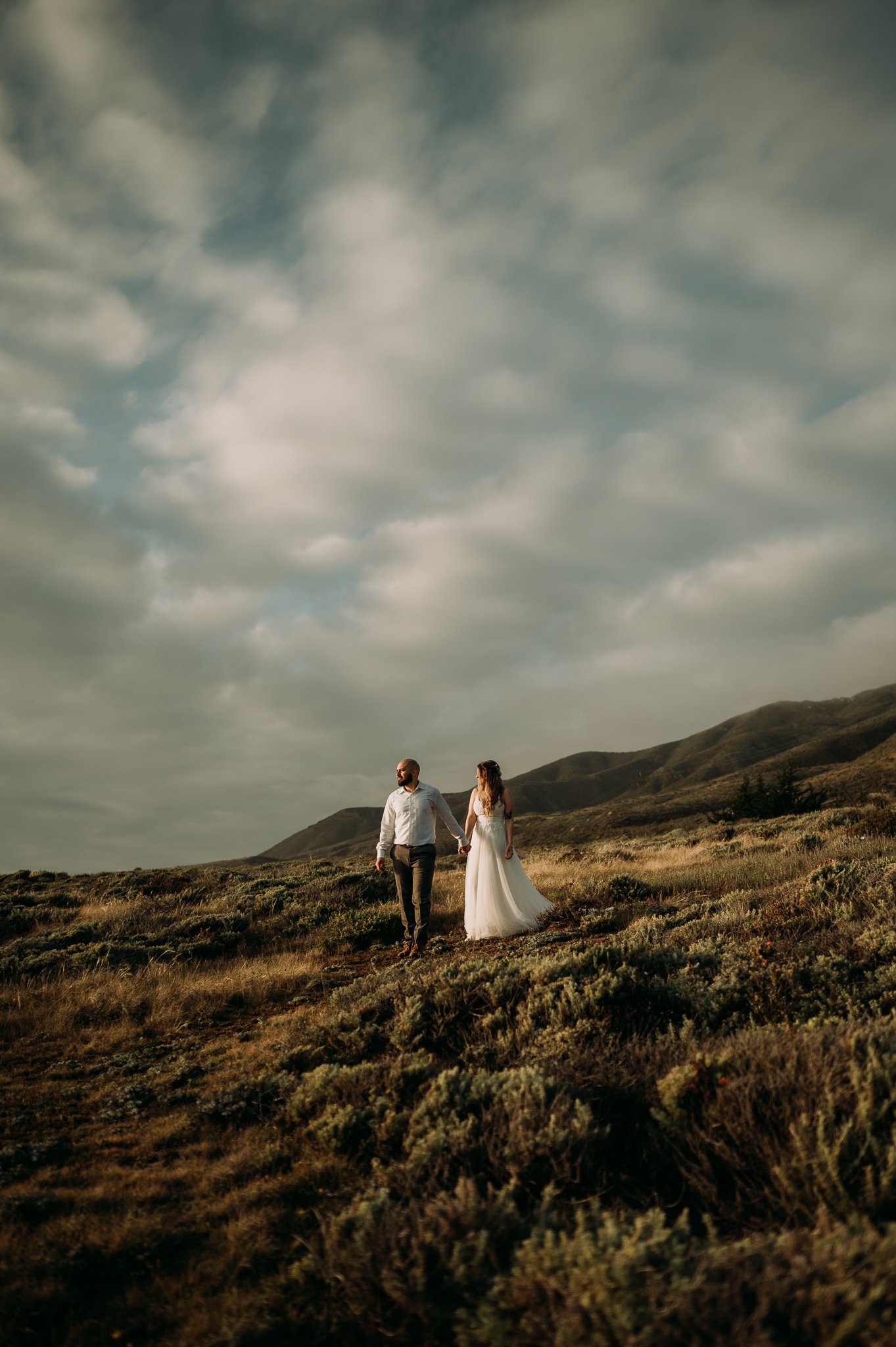 bride and groom standing in field with moody grey skies and hills in background gazing at Pacific Ocean Big Sur