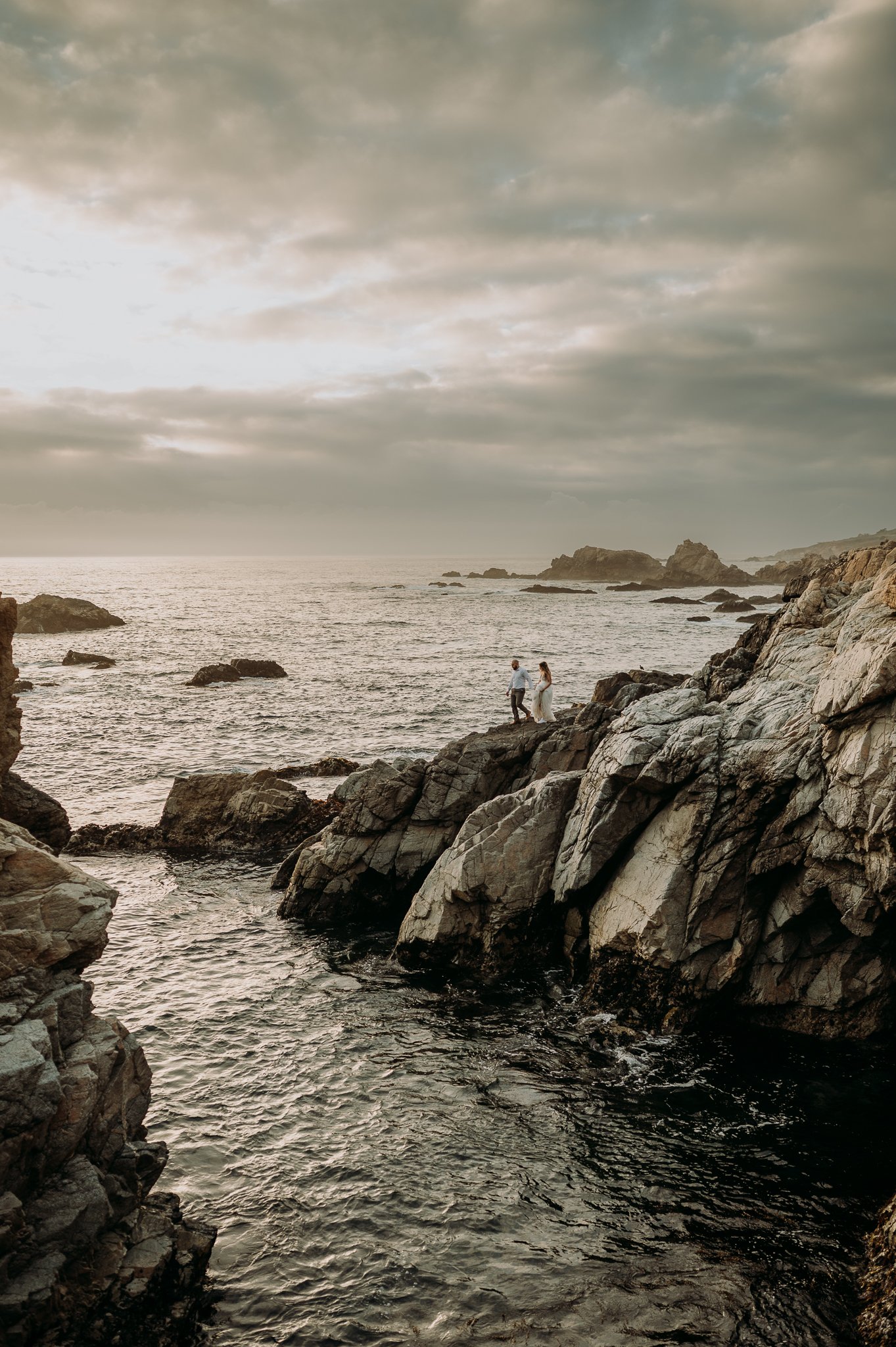 newly married couple walking out to edge of cliff holding hands in wedding dress and suit pacific ocean and moody grey skies in background 