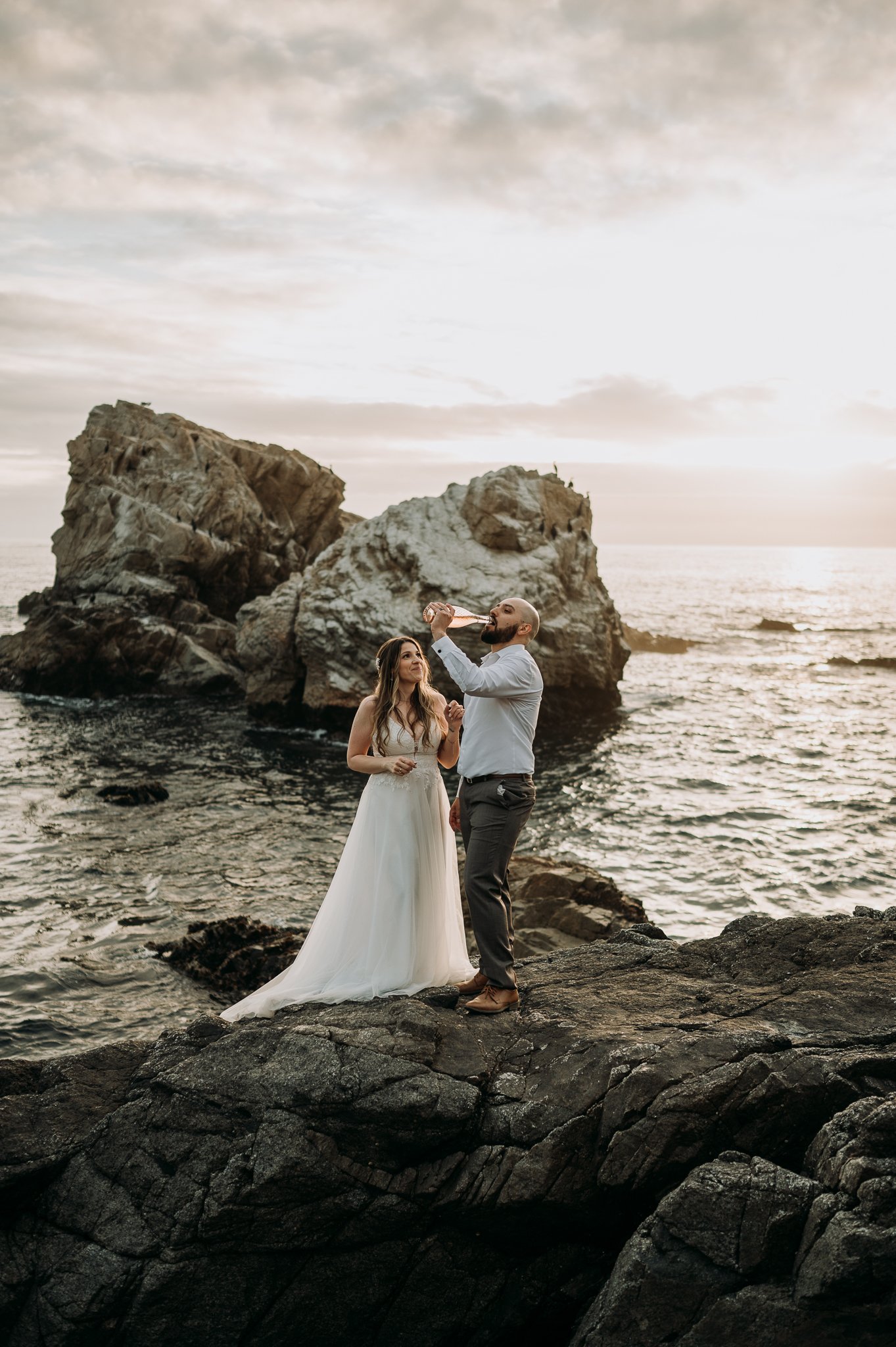 newly married couple celebrating elopement with champagne at edge of cliff in wedding dress and suit pacific ocean and moody grey skies in background 