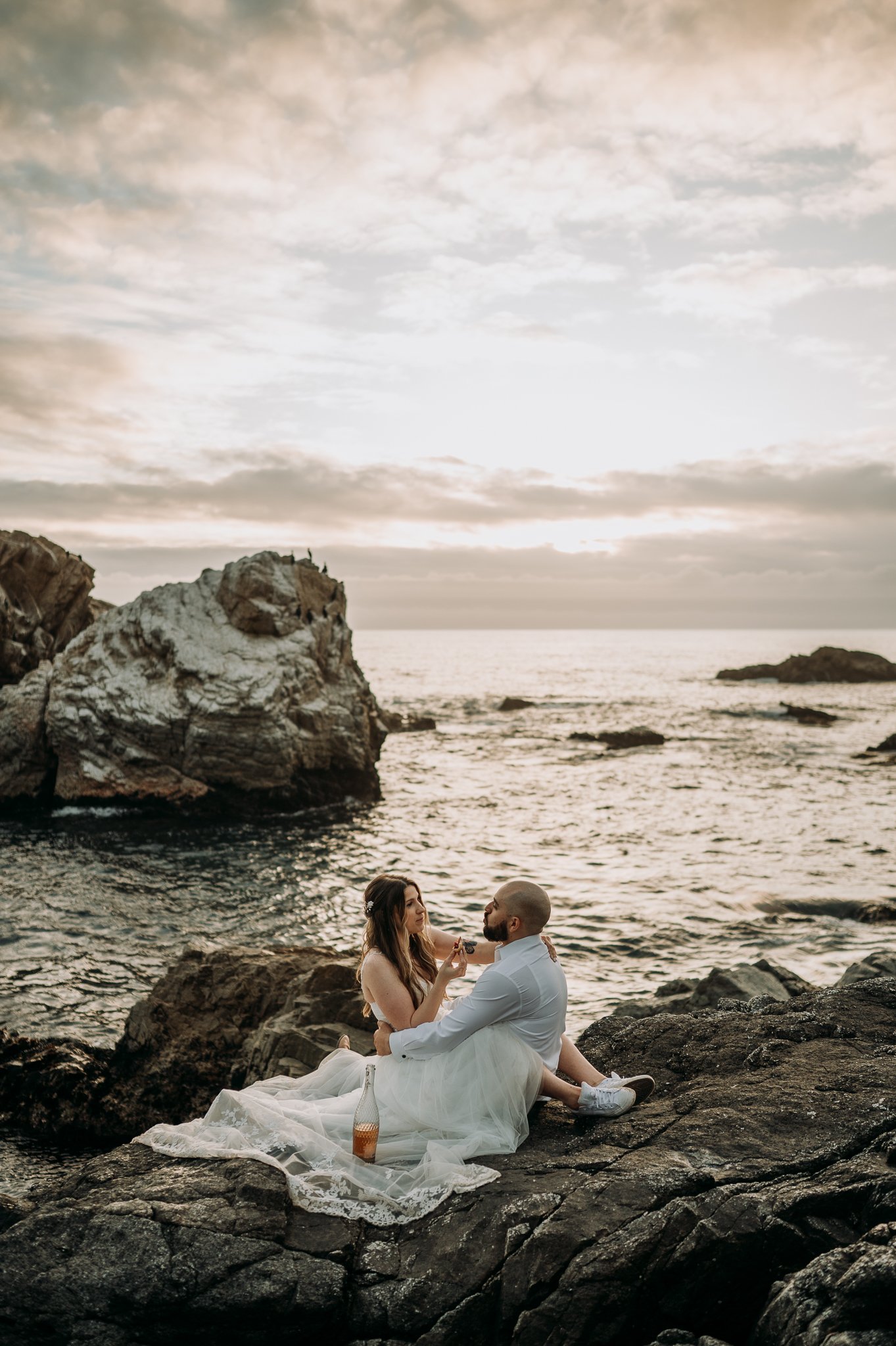 Couple sitting on cliff with Pacific Ocean in background arms intwind eating pie and drinking champagne
