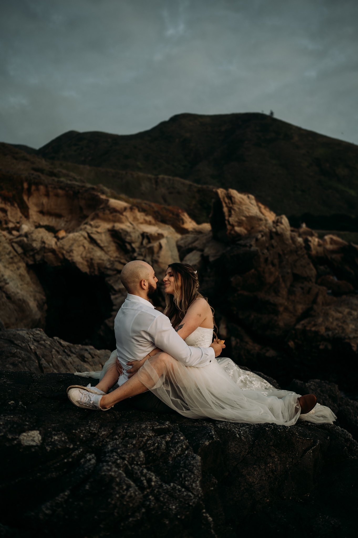 bride and groom sitting with legs intwind looking at each other with a smile on Big Sur coast