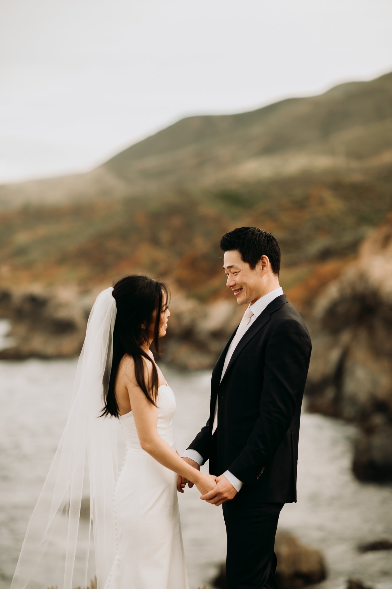 bride and groom standing on cliff above pacific ocean in Big Sur California holding hands looking at each other face to face