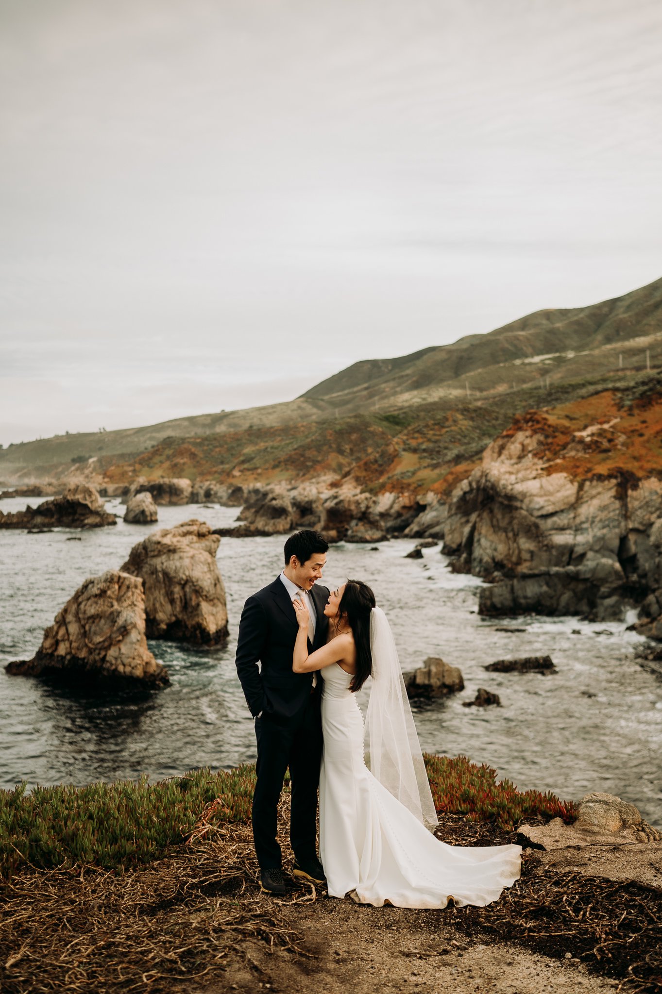 bride and groom standing on cliff above pacific ocean in Big Sur California looking at each other face to face