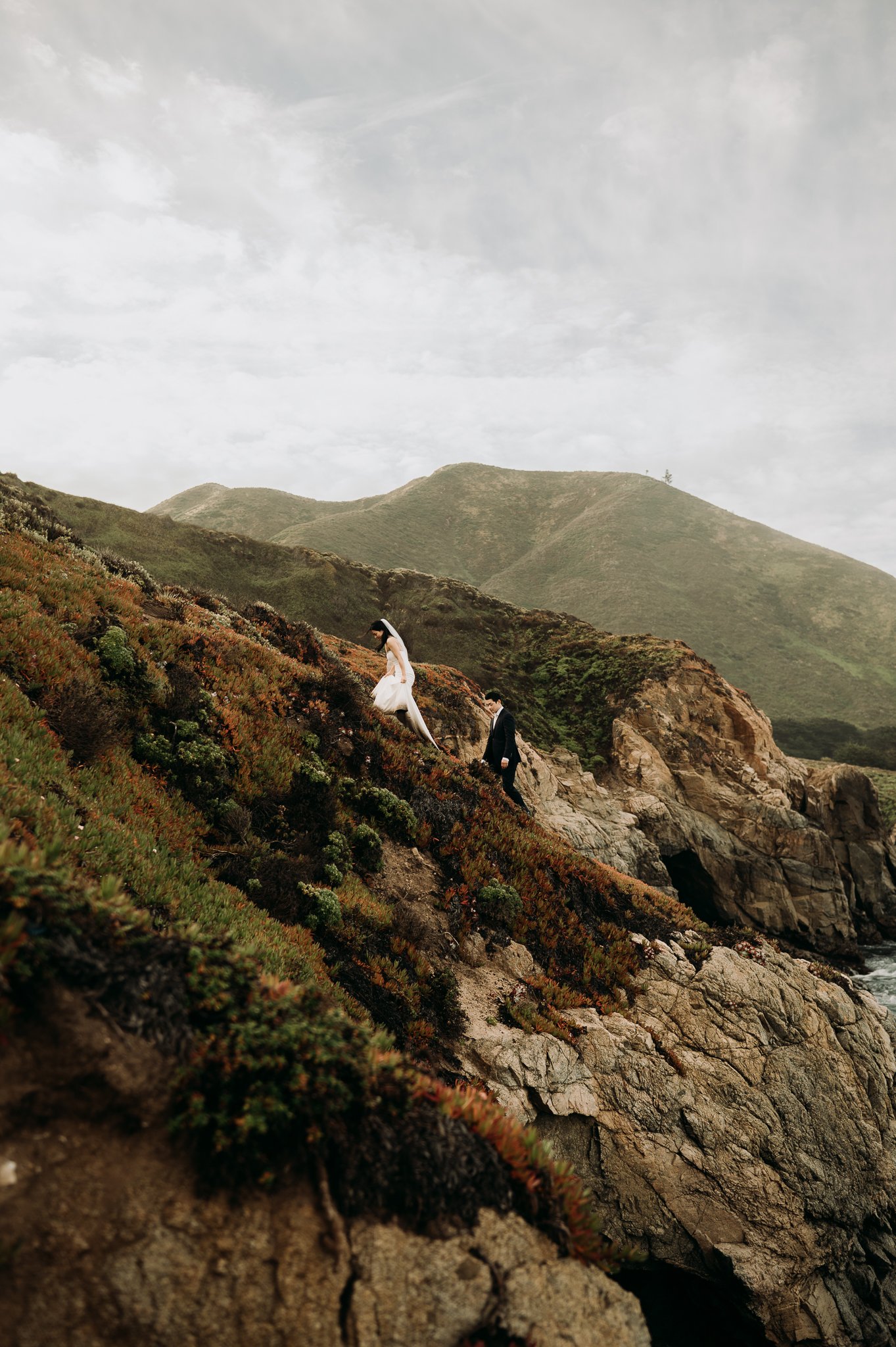 Big Sur Elopement couple walk up trail with hills and moody grey skies in background