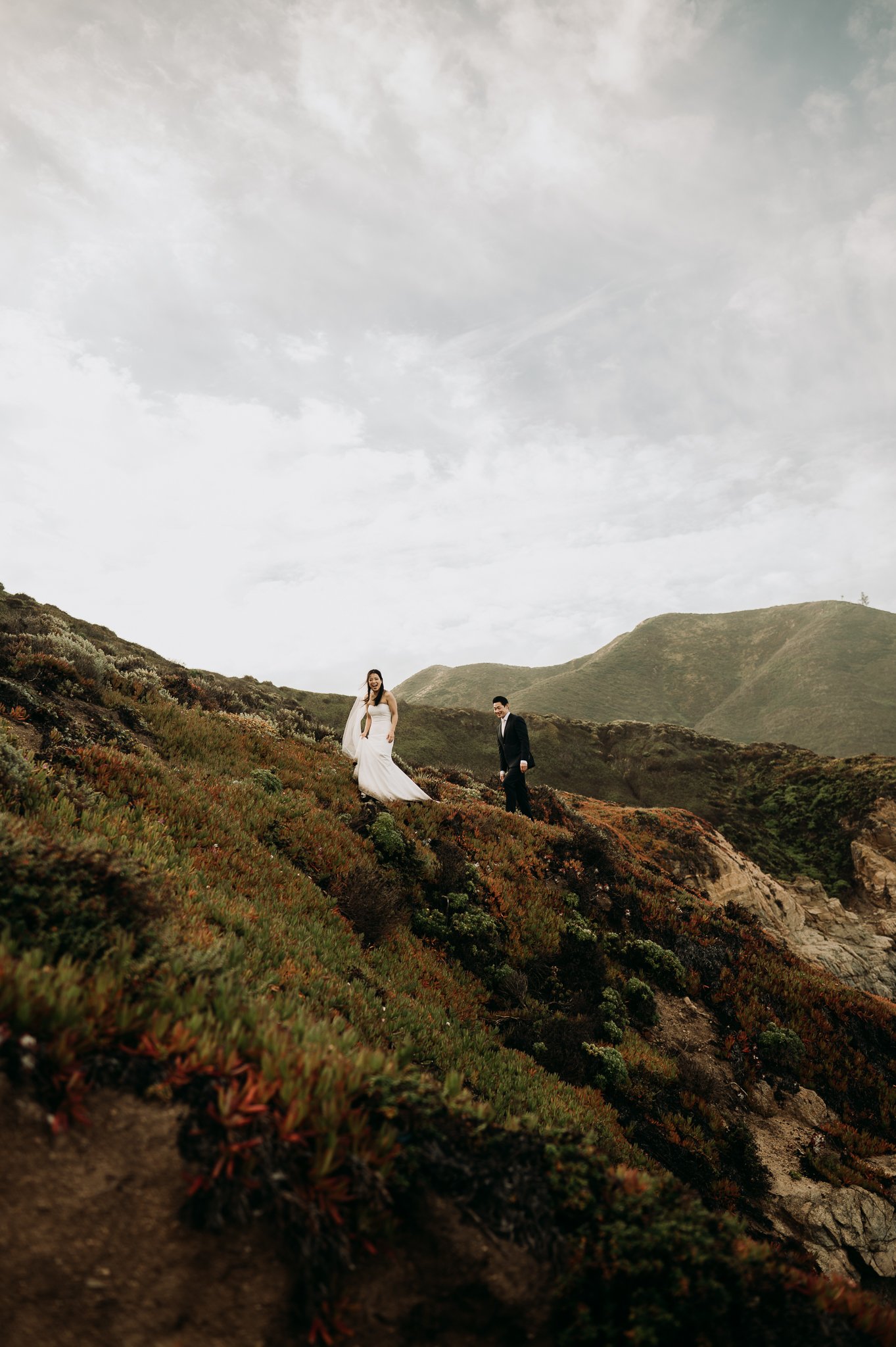 Big Sur Elopement couple walk up trail and smile at photographer with hills and moody grey skies in background
