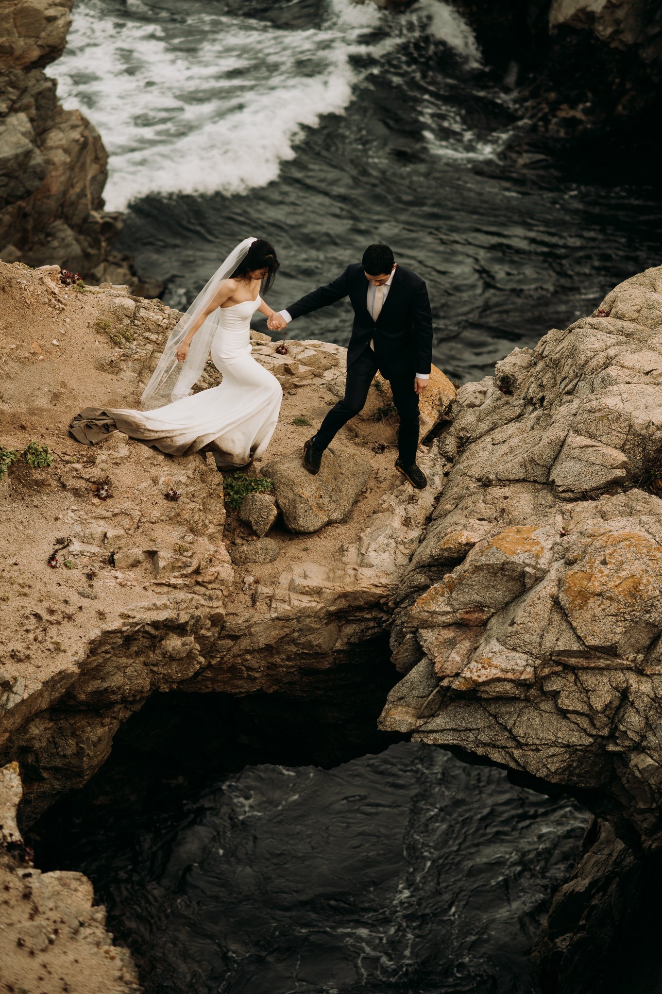 Groom helps bride in wedding dress to a cliffside spot to pose for photo