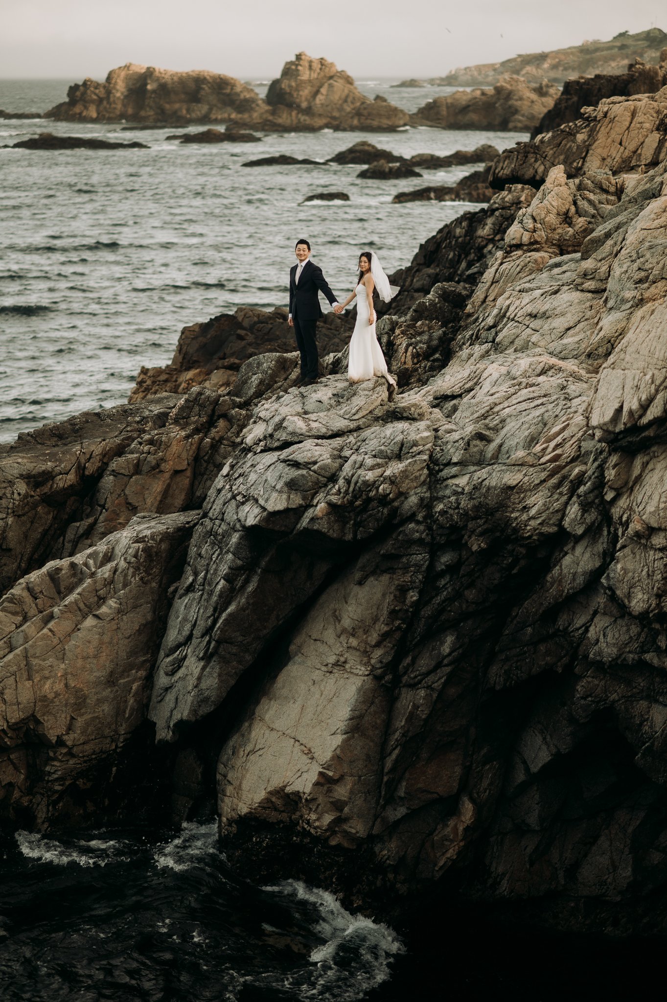 Bride and groom posing for photo on cliff with Pacific Ocean in background
