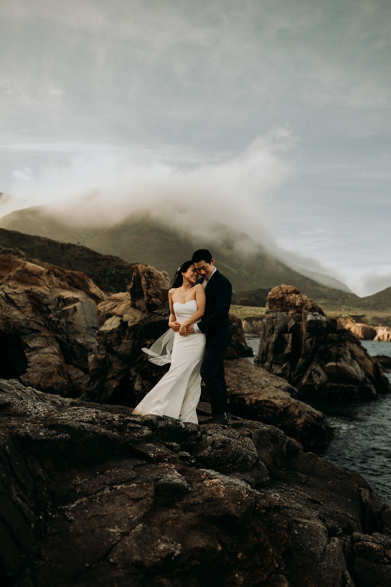 Bride and groom embracing with bride in front of groom on cliff with Pacific Ocean in background Big Sur Elopement