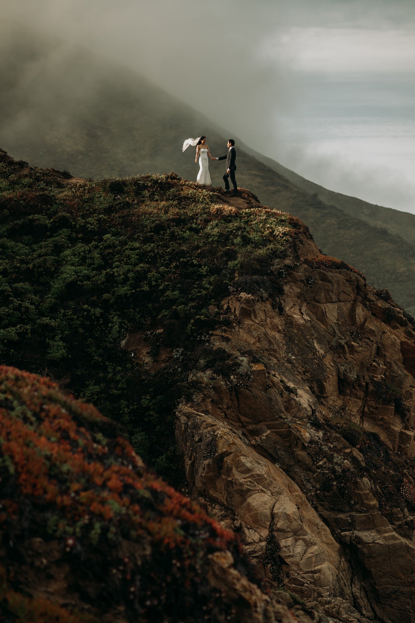 Elopement couple in wedding attire hugging on cliff in with ocean and fog drenched hills Big Sur California