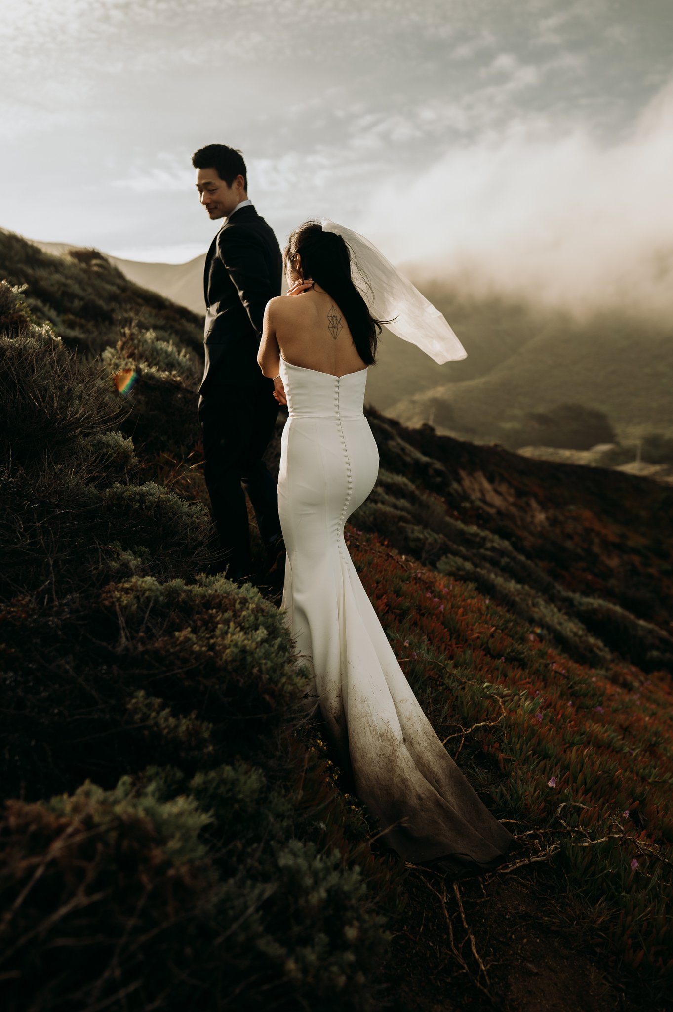 Elopement couple in wedding attire walking up trail with fog drenched hills Big Sur California