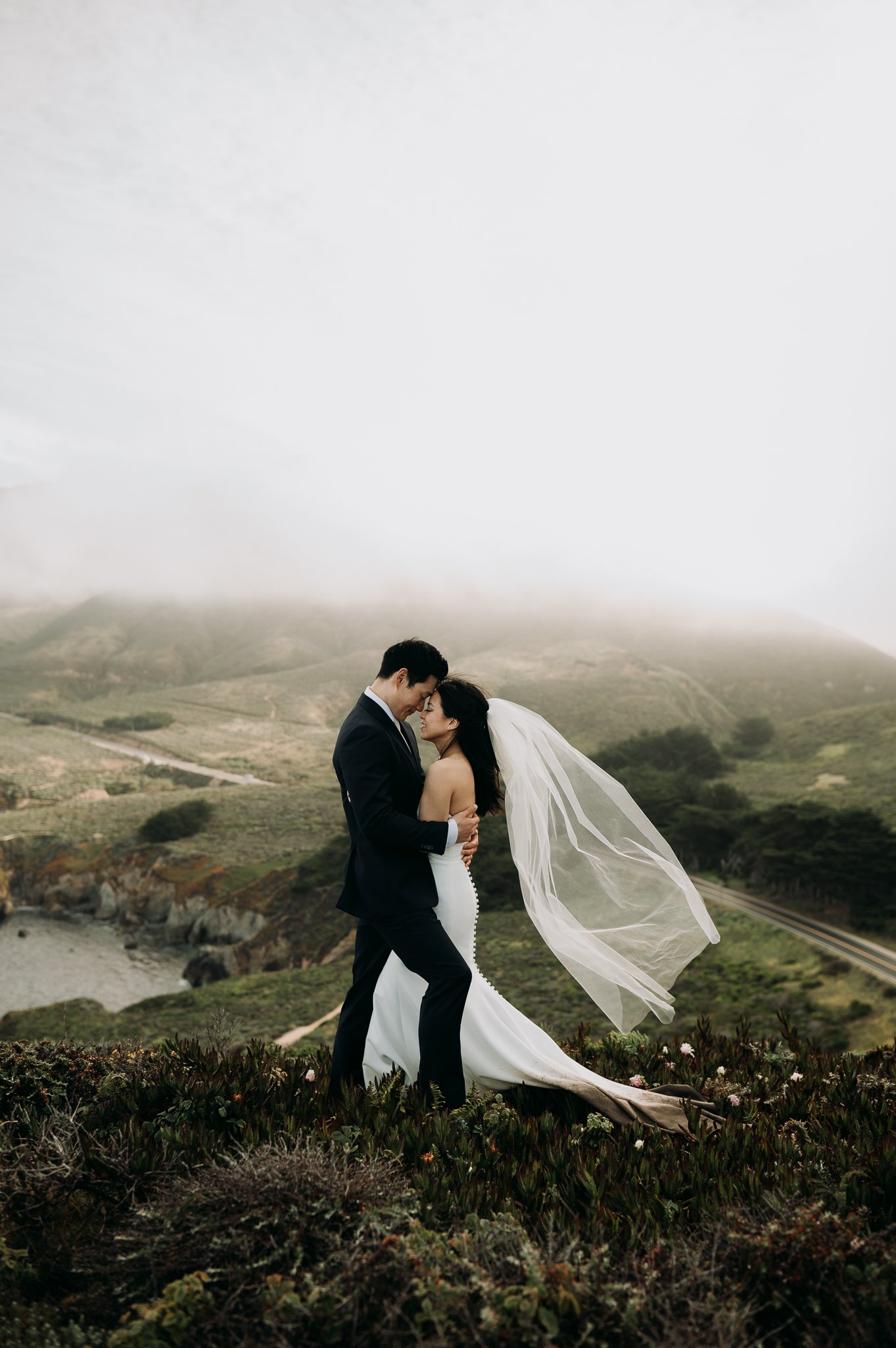 Elopement couple in wedding attire hugging on cliff in with ocean and fog drenched hills Big Sur California