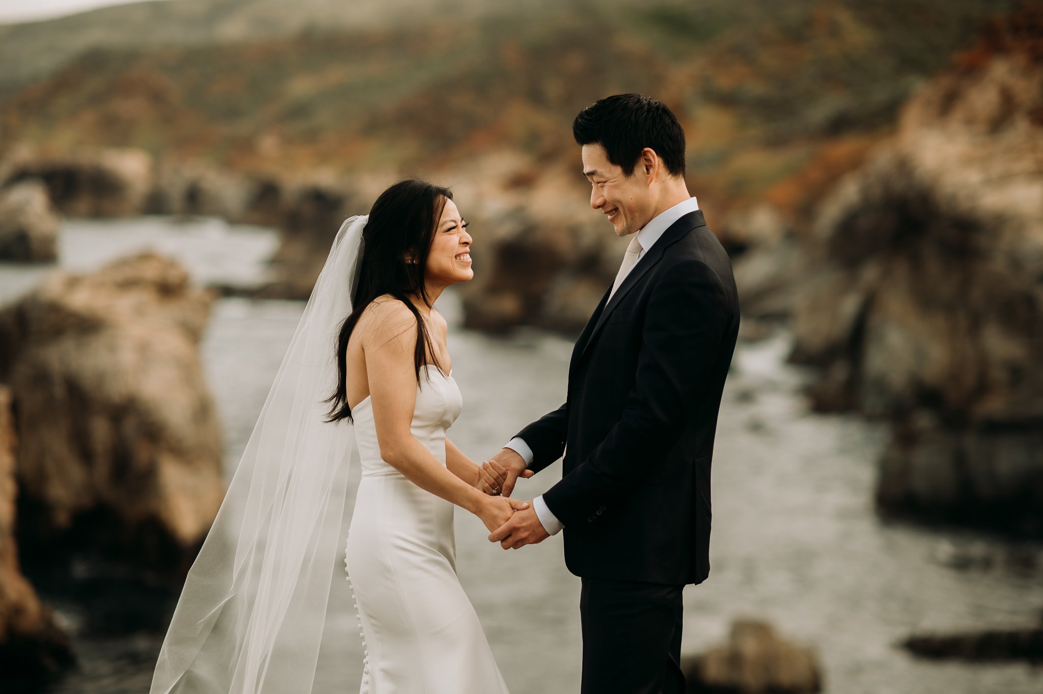 bride and groom standing on cliff above pacific ocean in Big Sur California holding hands smiling