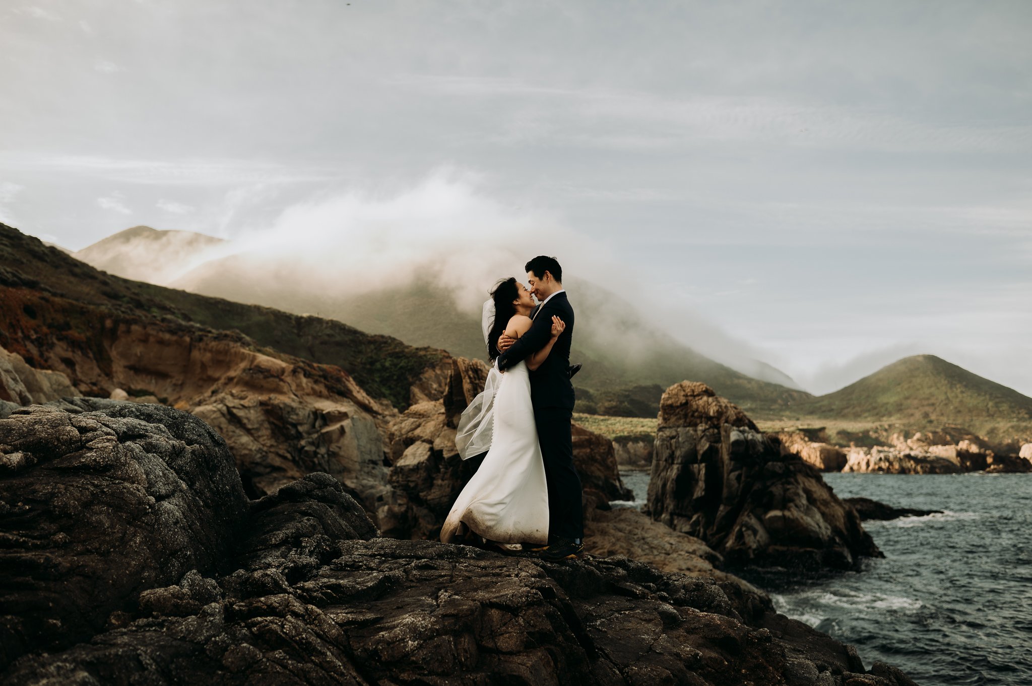 Elopement couple in wedding attire hugging on cliff in with ocean and fog drenched hills Big Sur California