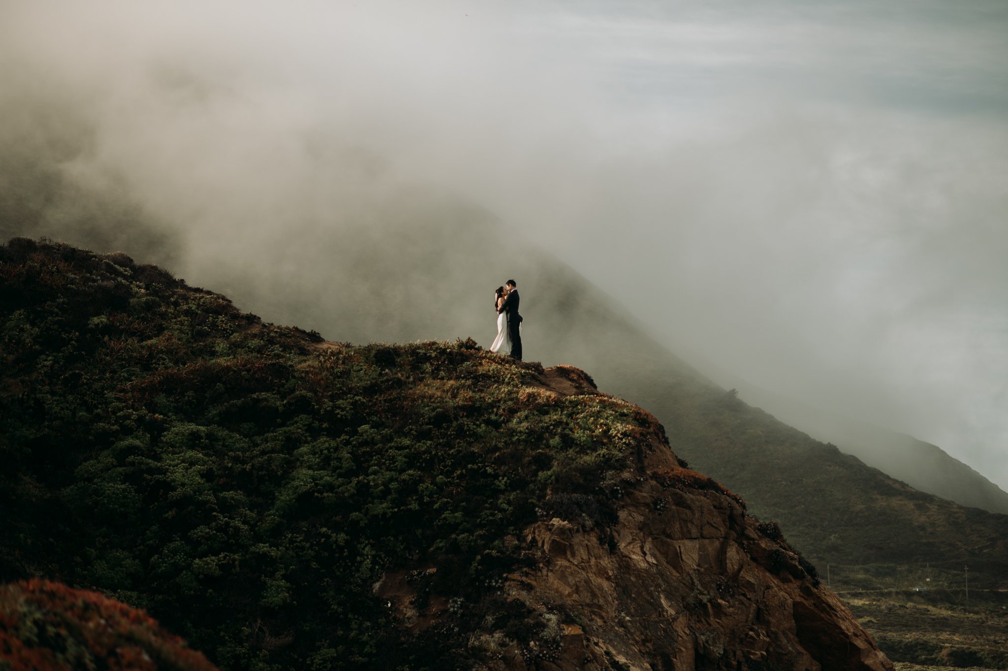 Elopement couple in wedding attire hugging on cliff in with ocean and fog drenched hills Big Sur California