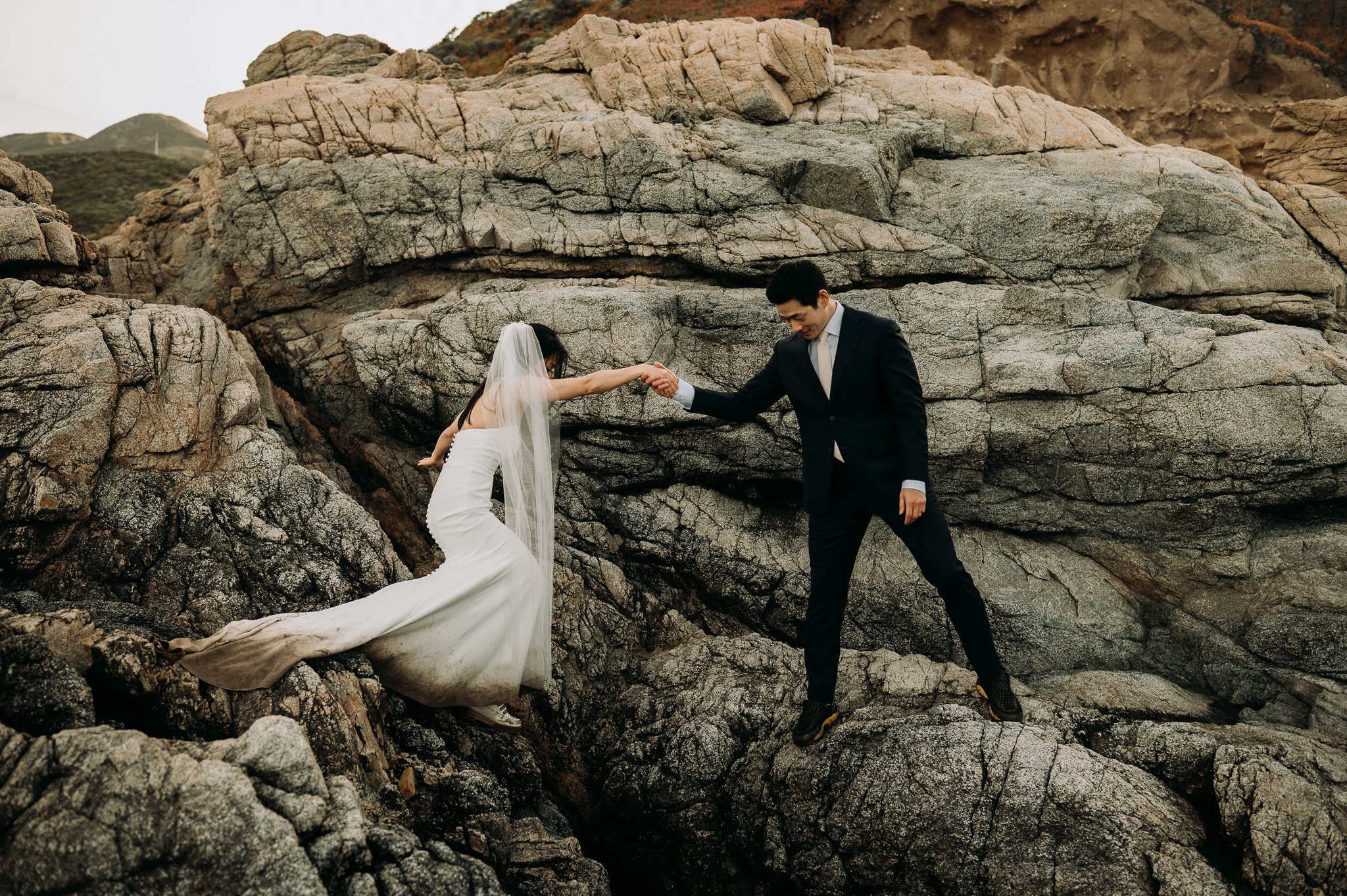 Groom helping bride adventuring cliffside Big Sur California 