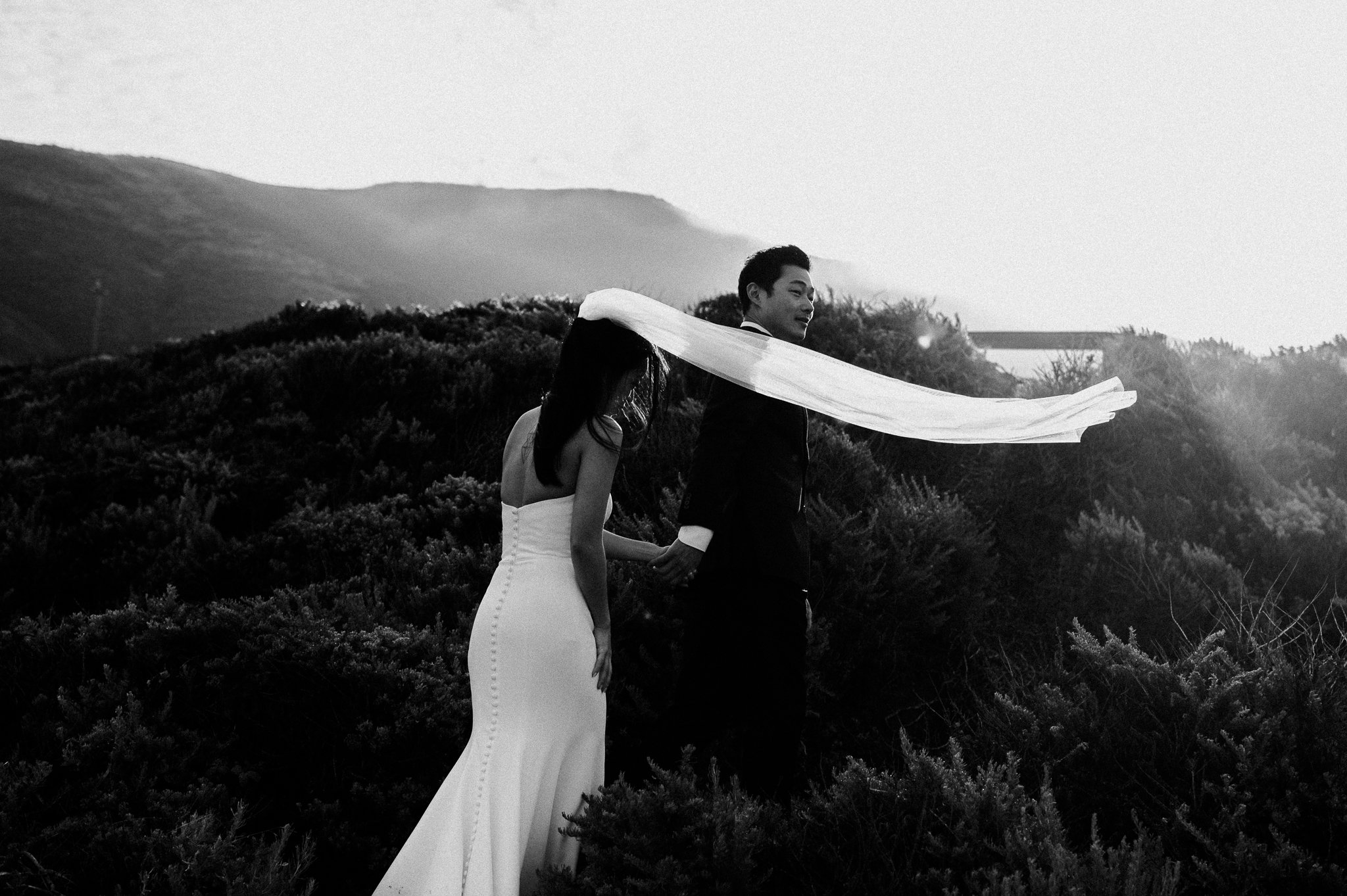 Black and white photo bride and groom walking on trail in wedding attire brides vail blowing in wind