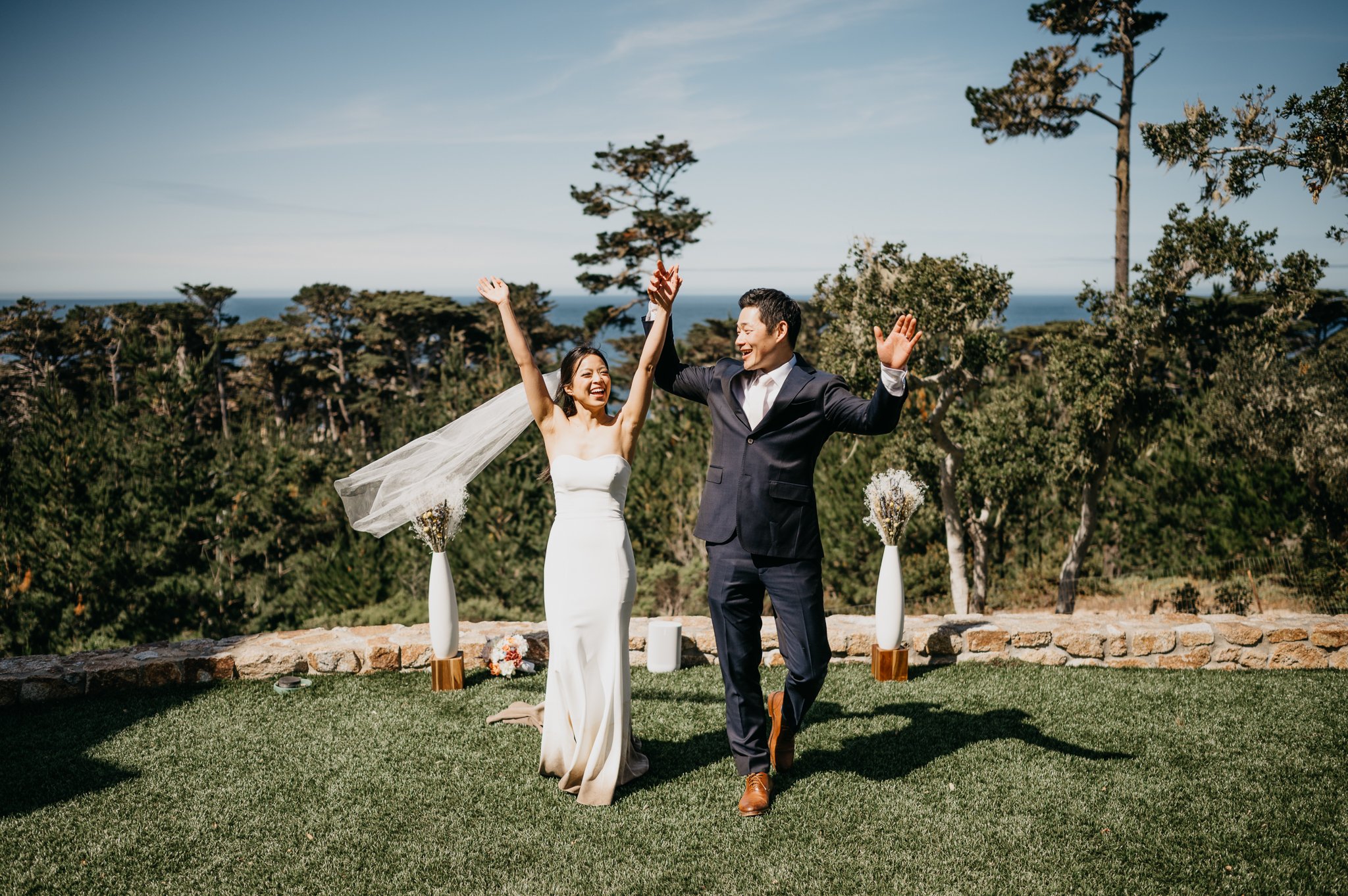 Bride and groom on grassy area with cypress tress in background with both hands in air celebrating their wedding Pebble Beach California